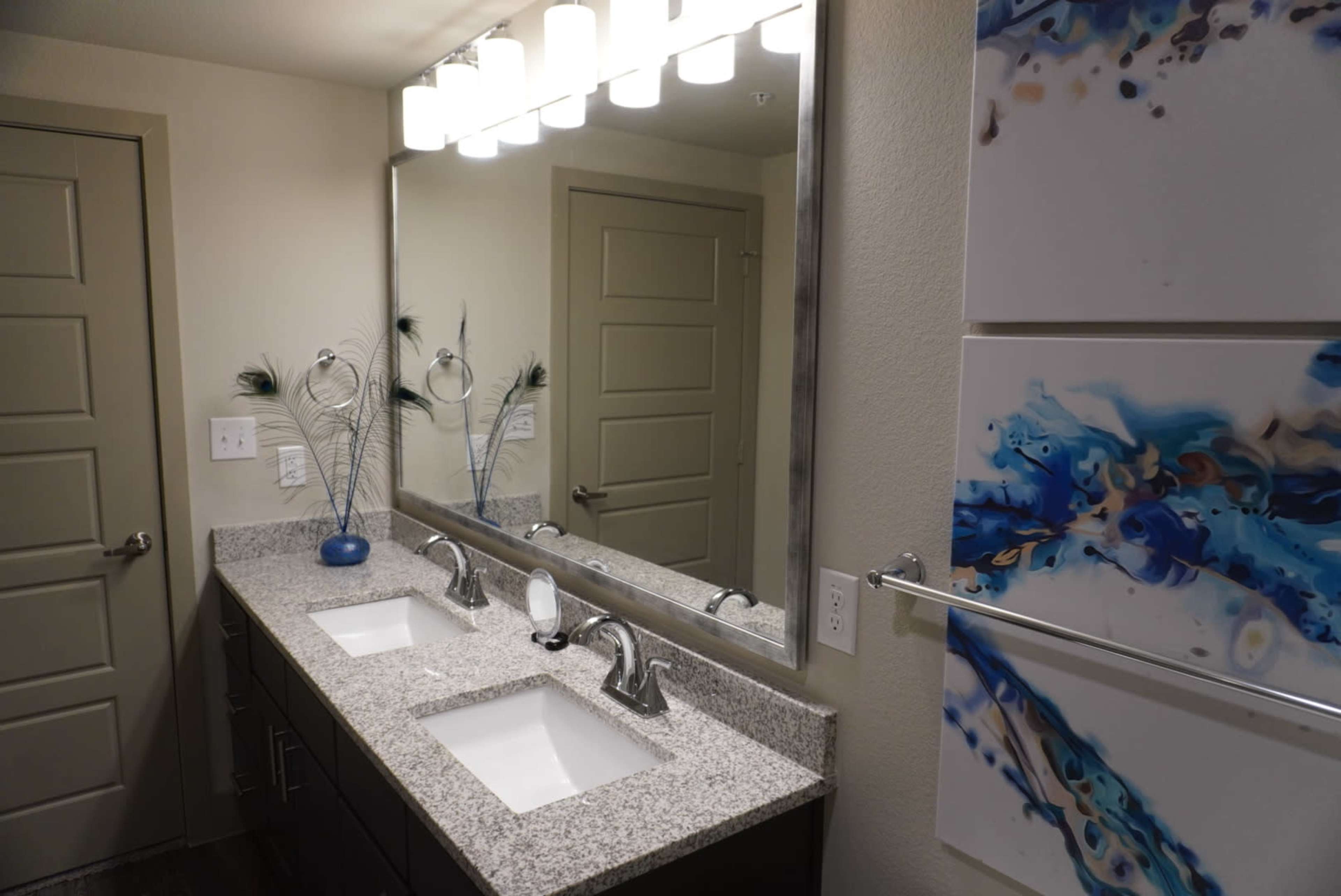 A modern bathroom features a double sink vanity with a granite countertop, a large mirror, and decorative artwork on the walls.
