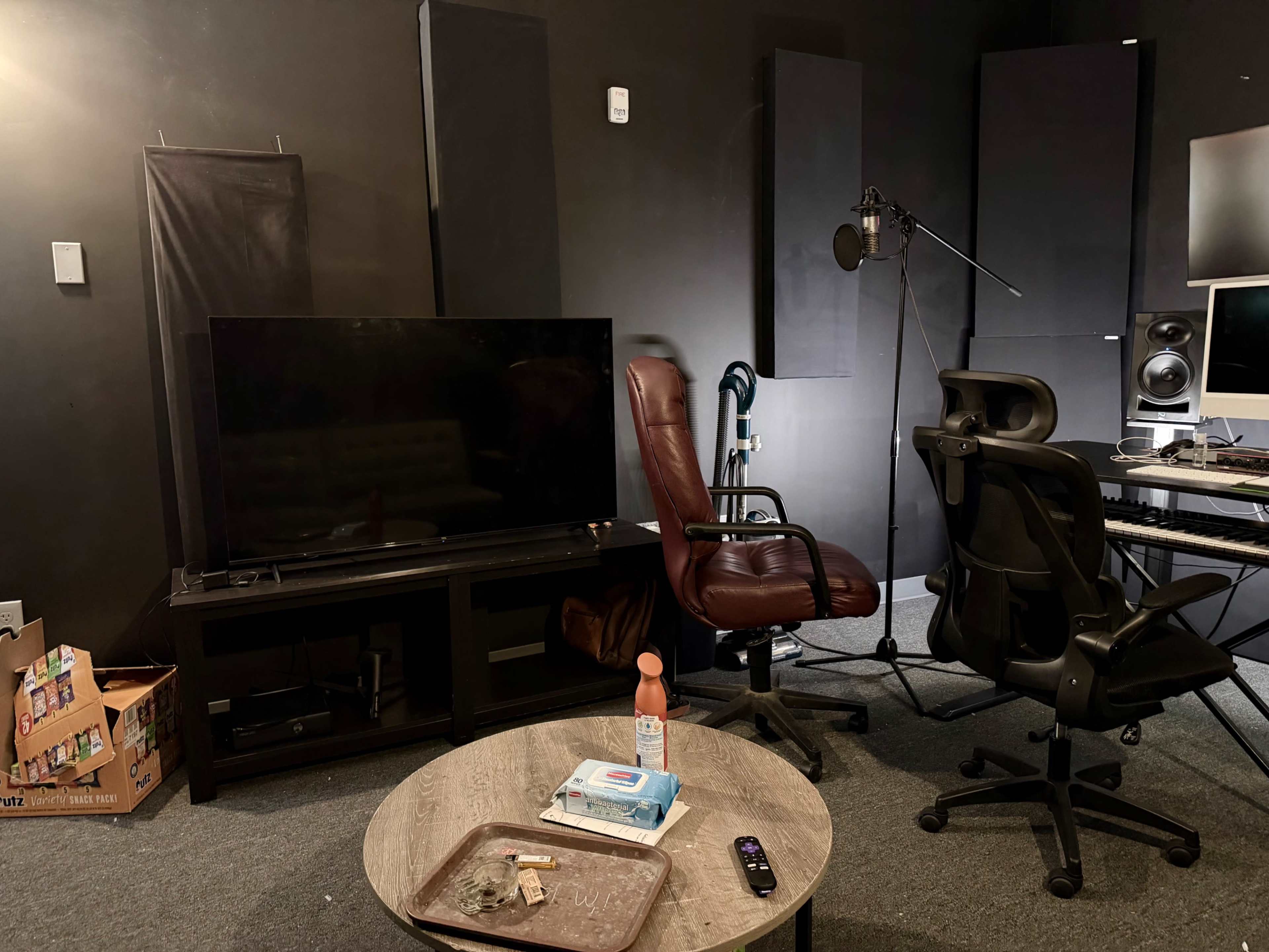 The image shows a recording studio setup with a large television, two chairs, a microphone on a stand, and various audio equipment against dark wall panels.