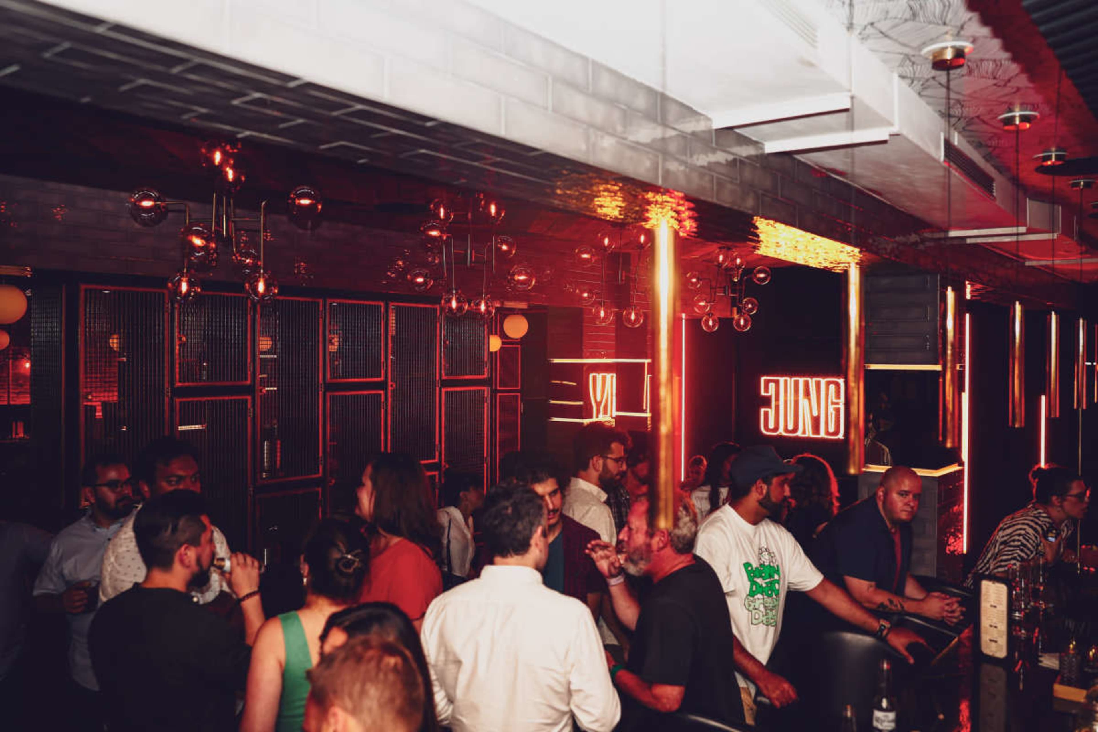 A crowded bar scene features patrons socializing under dim red lighting with neon signs in the background.