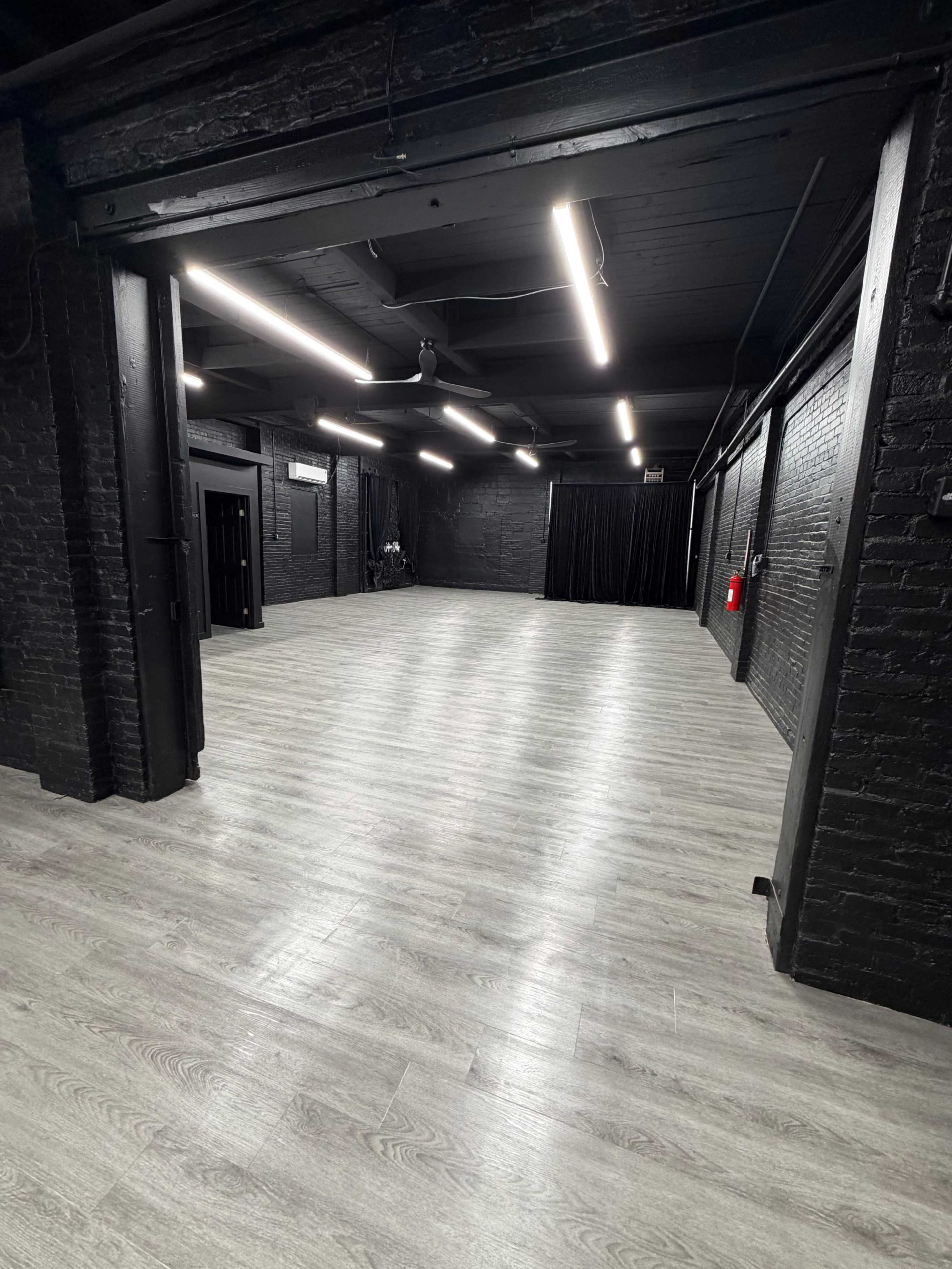 The image shows a spacious, empty room with black brick walls and gray laminate flooring, featuring overhead fluorescent lights.