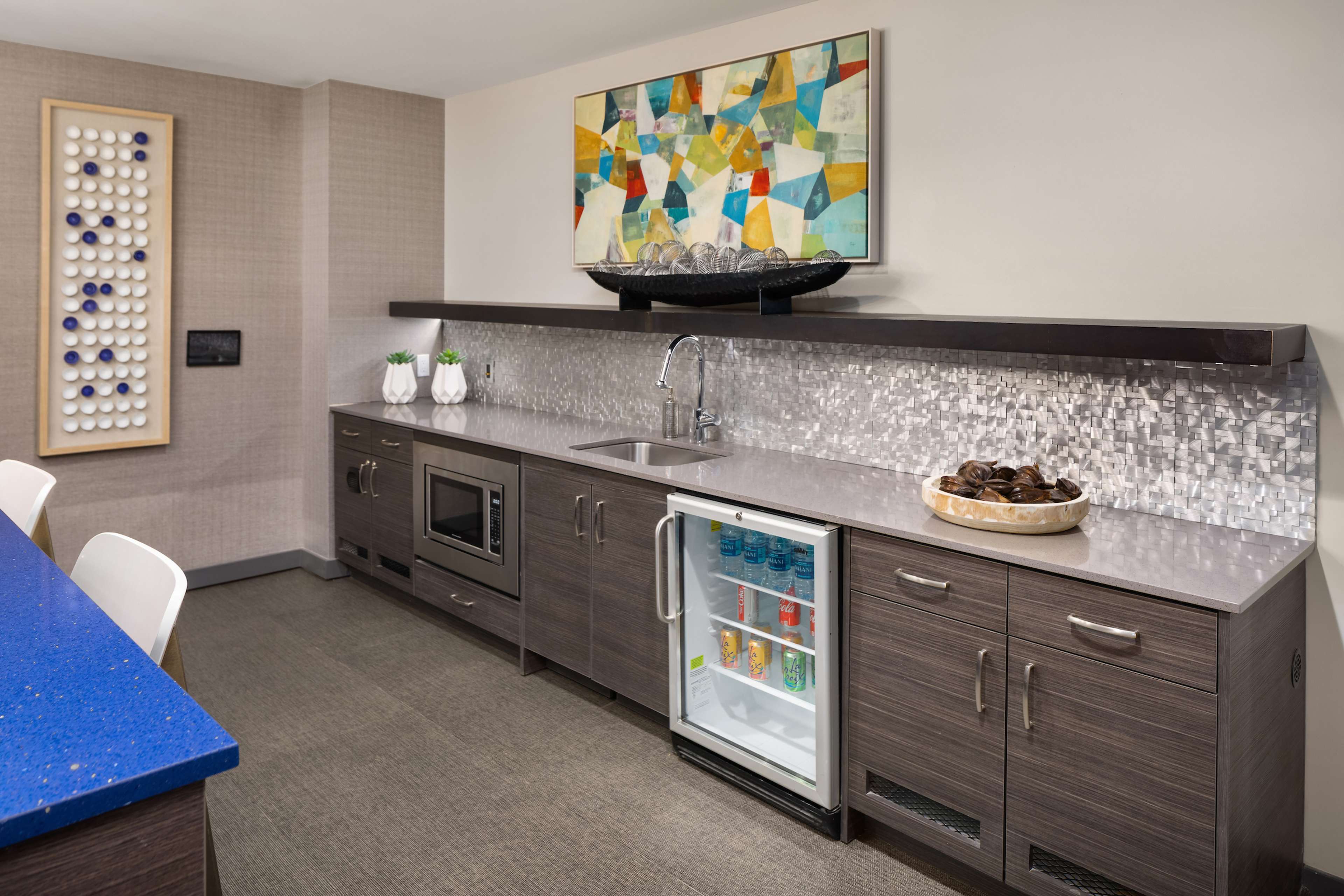 A modern kitchen with a blue countertop, stainless steel appliances, a mosaic backsplash, and a small refrigerator displaying various drinks.