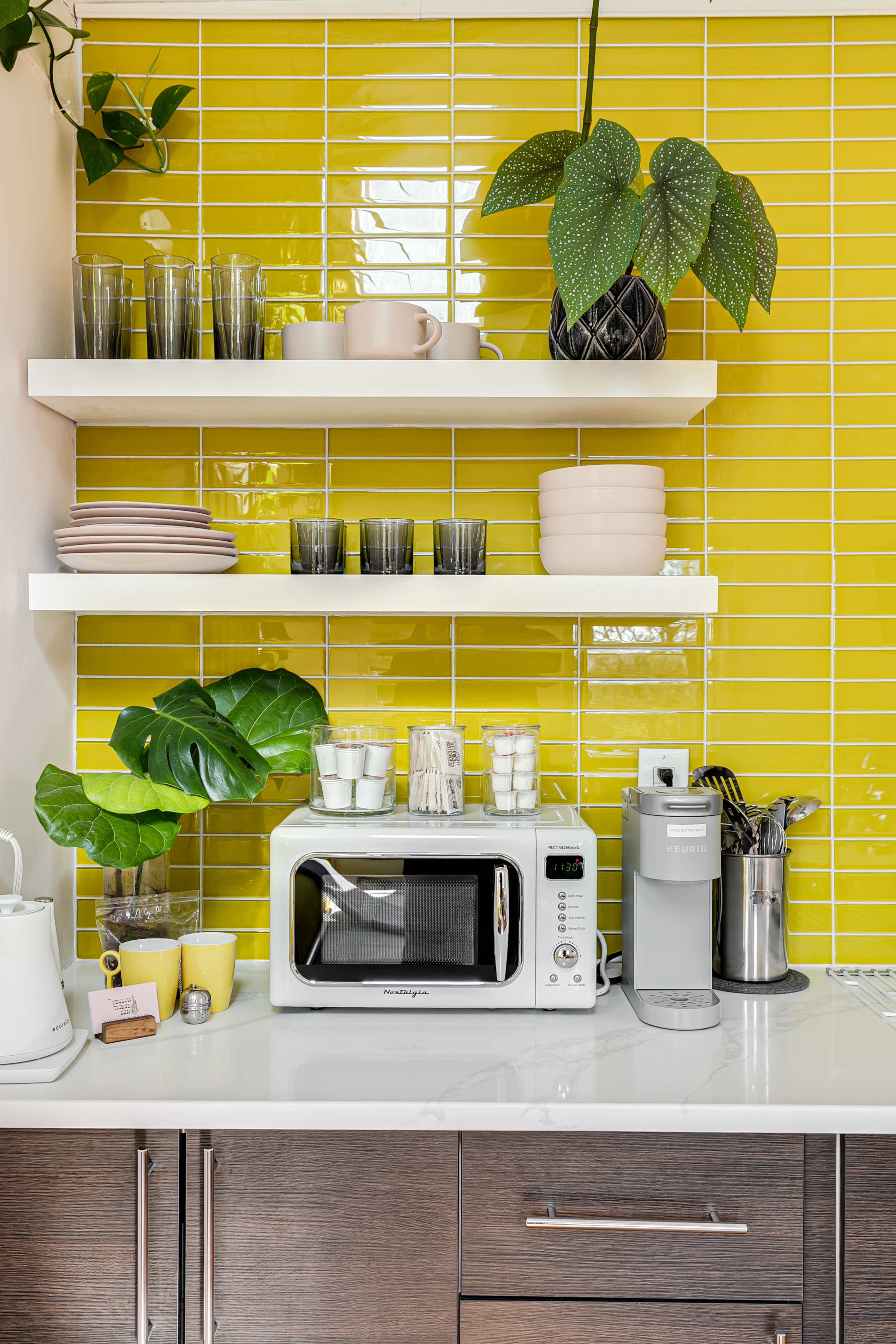 A modern kitchen features a bright yellow tiled wall, two shelves with various dishware and glassware, a countertop microwave, and decorative plants.