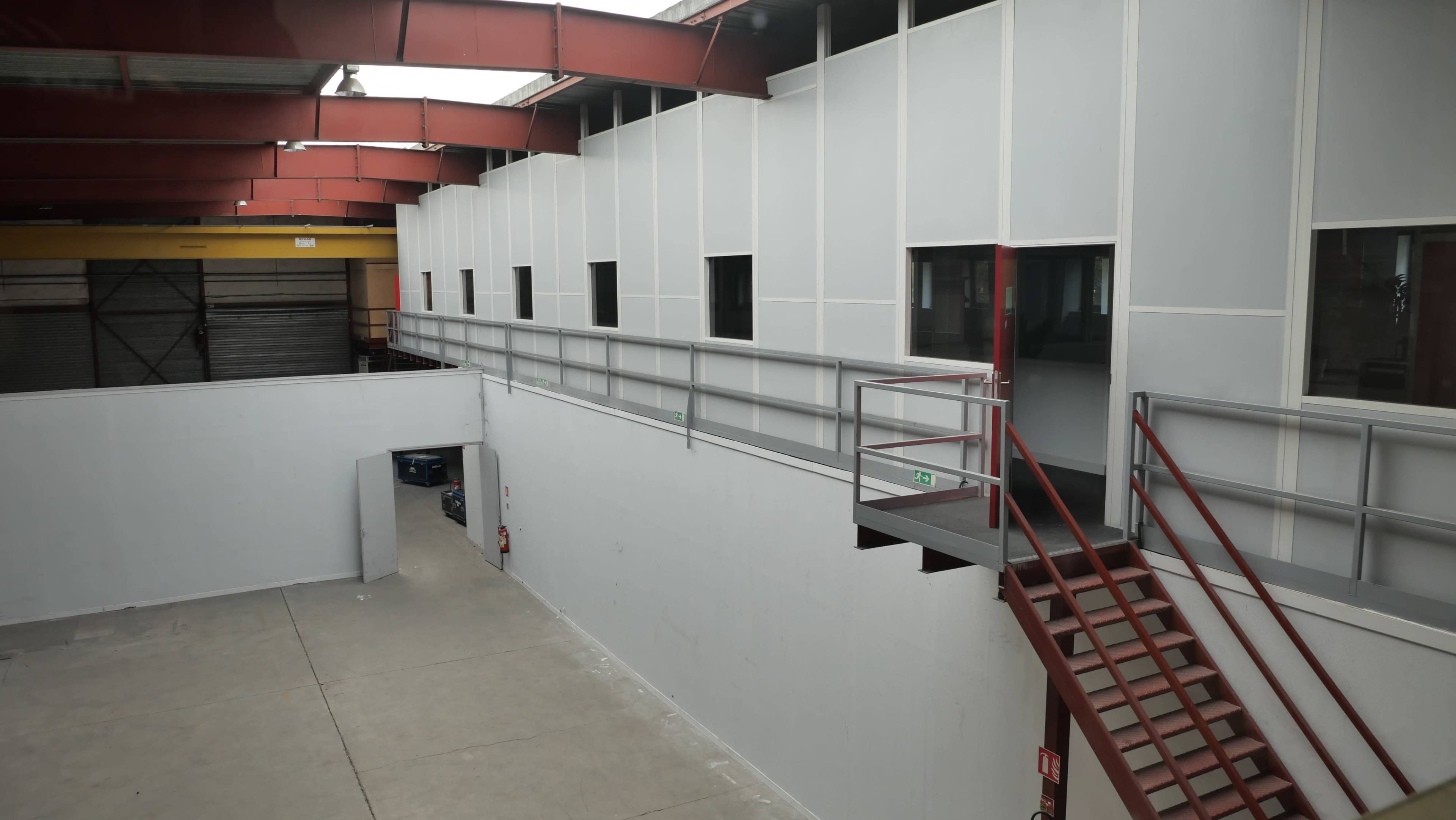 A spacious industrial interior with white walls, large windows, and a red metal staircase leading to an upper level.