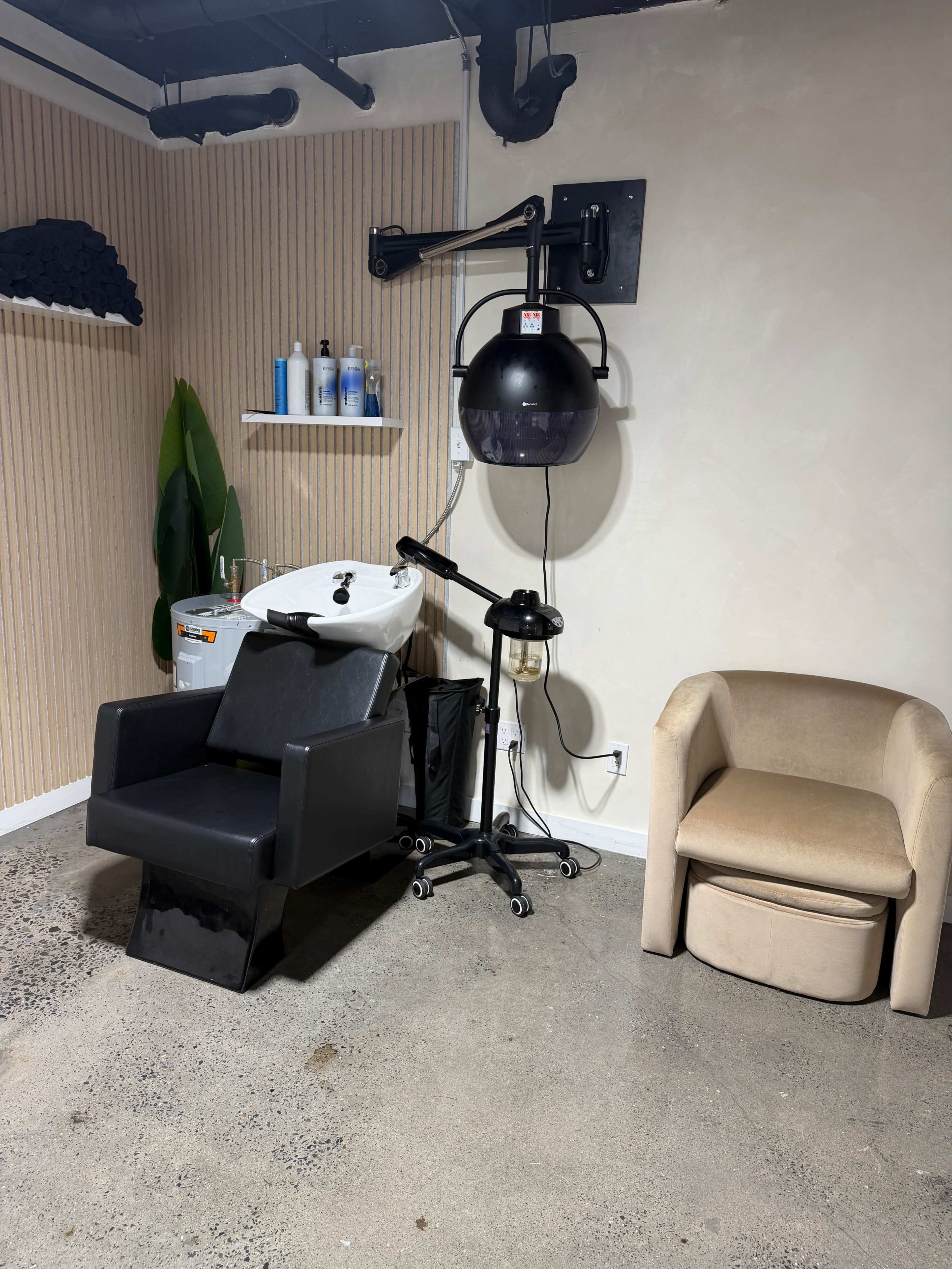 The image shows a hair salon corner with a black shampoo chair, a hooded dryer on wheels, and a light-colored accent chair against a textured wall.