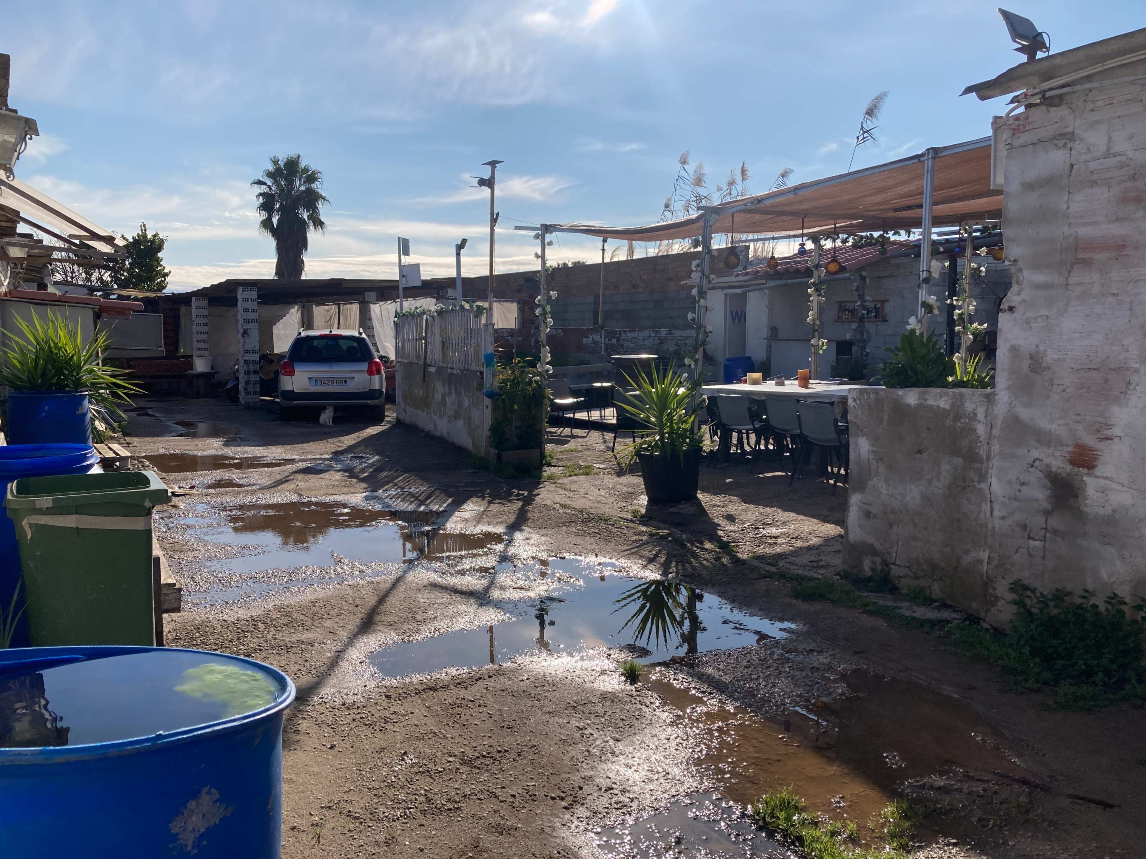 The image depicts a gravel pathway lined with potted plants and tables, featuring puddles of water on the ground and a parked car in the background.