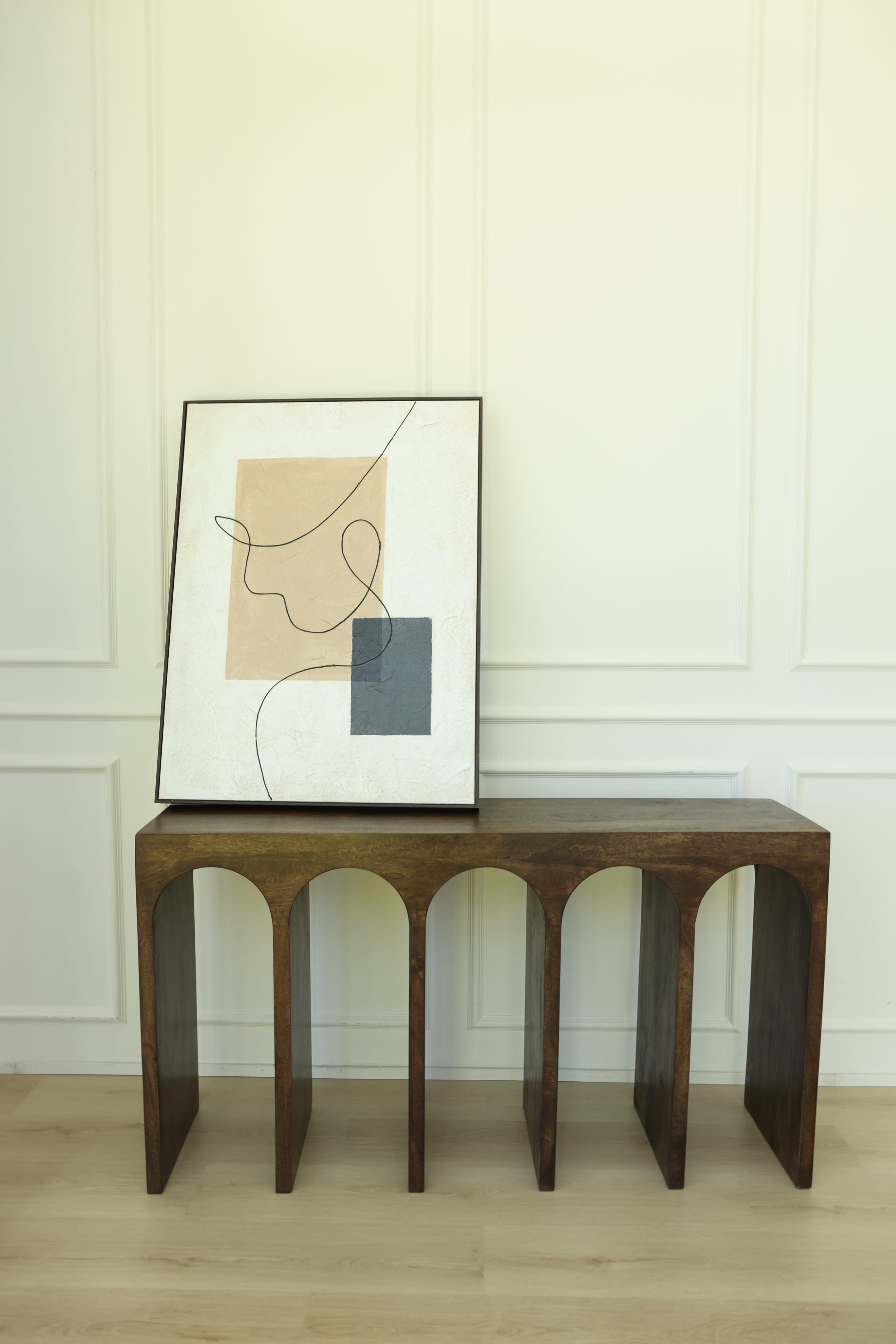 A wooden console table featuring arched cutouts holds a framed abstract artwork against a light-colored wall.