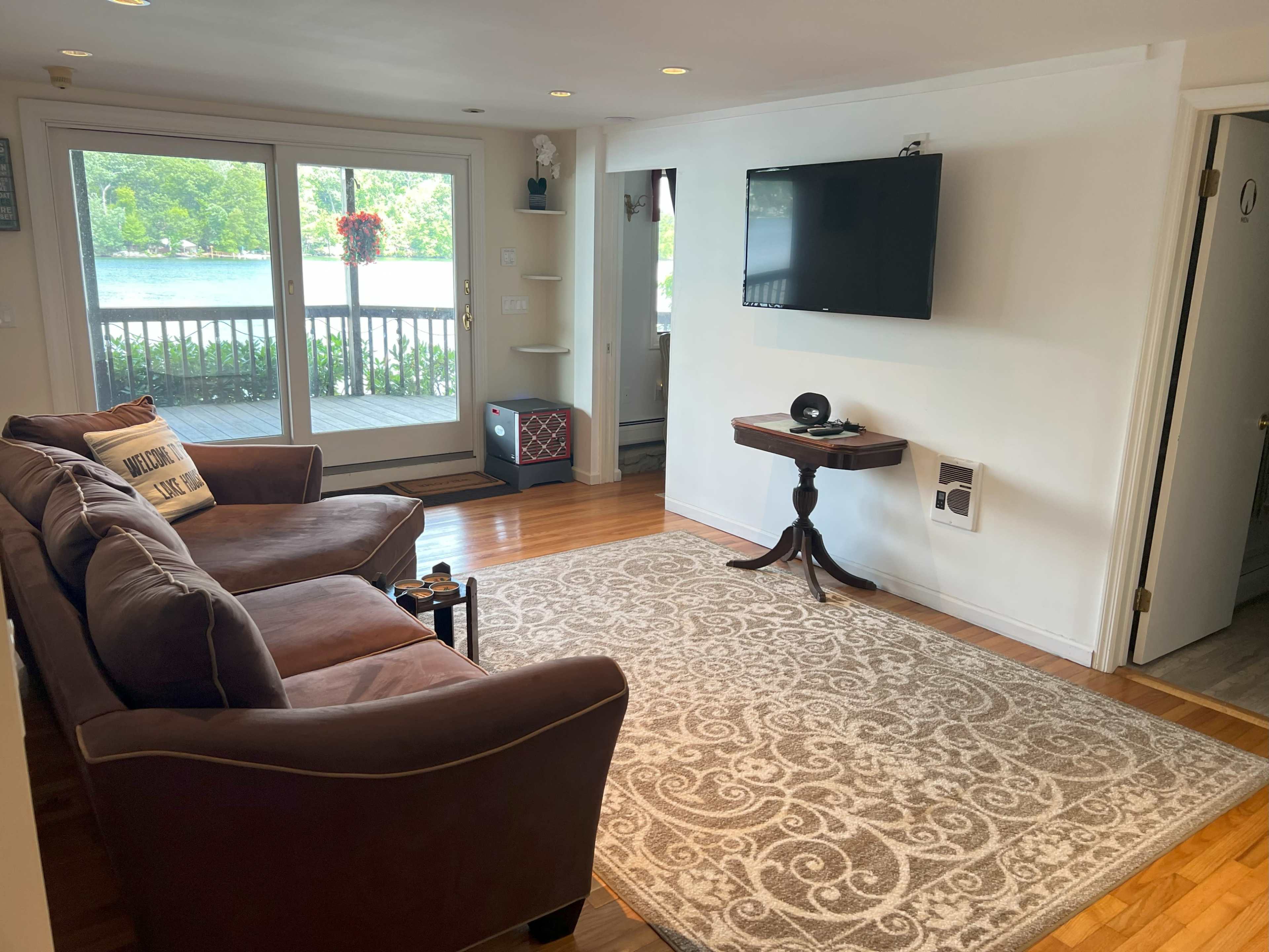 The image shows a living room furnished with a brown couch, a coffee table, and a wall-mounted television, overlooking a patio with a view of a body of water.