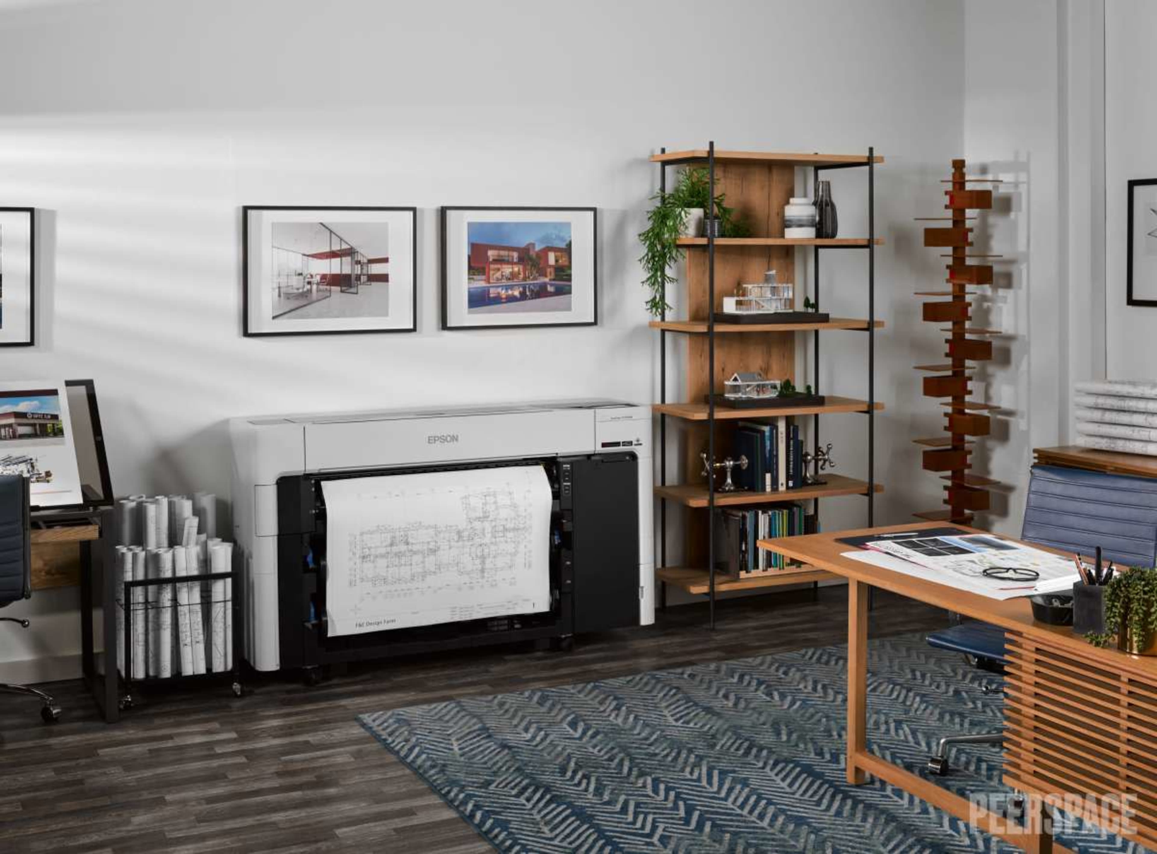 An office space features an Epson printer alongside shelves filled with books and decorative items, with blueprints rolled up on the floor and artwork displayed on the walls.