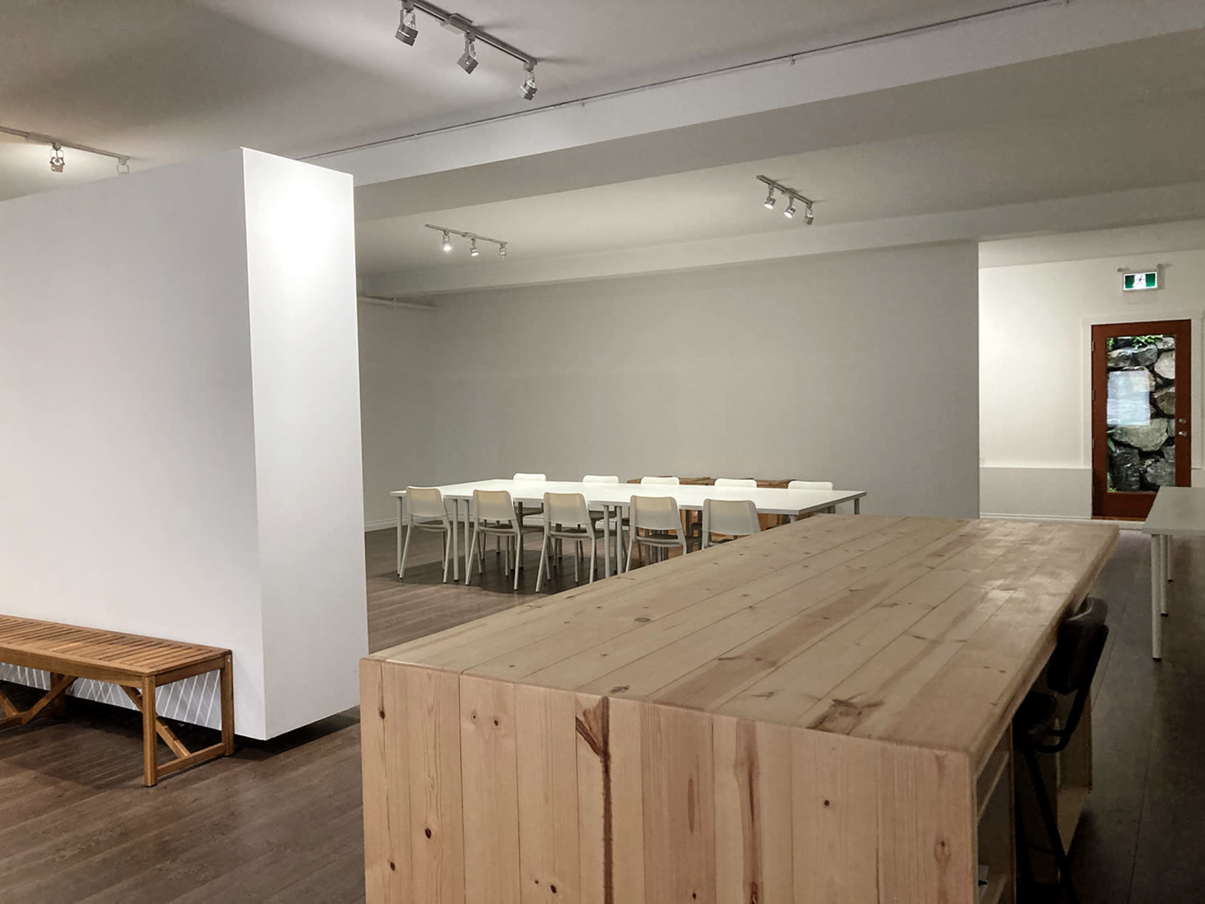A minimalist interior space with a long table and chairs arranged for a meeting, a wooden bench against one wall, and a counter made of light-colored wood.