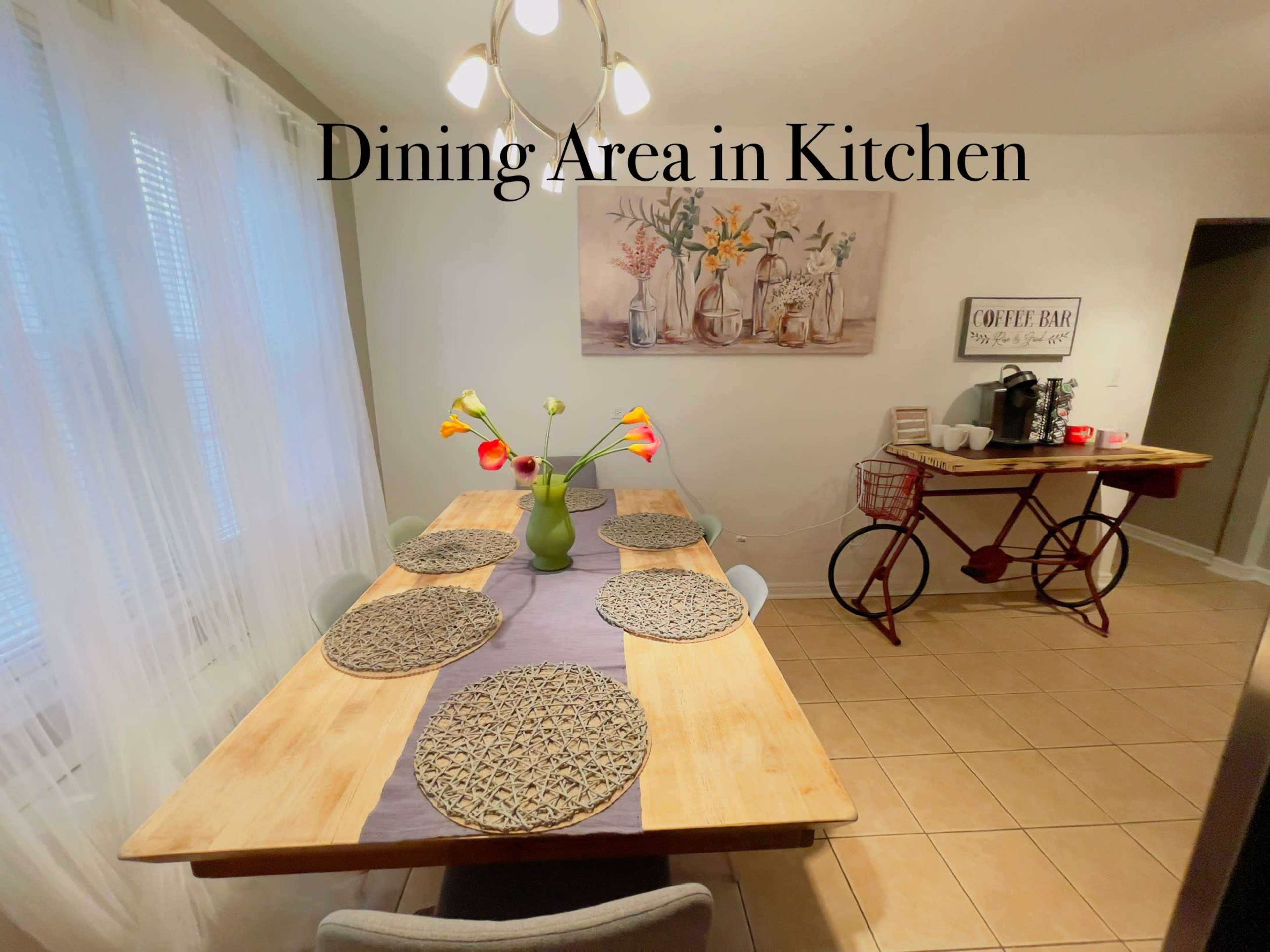 A dining table with six chairs is set in a kitchen space, decorated with a vase of flowers and a coffee station nearby.