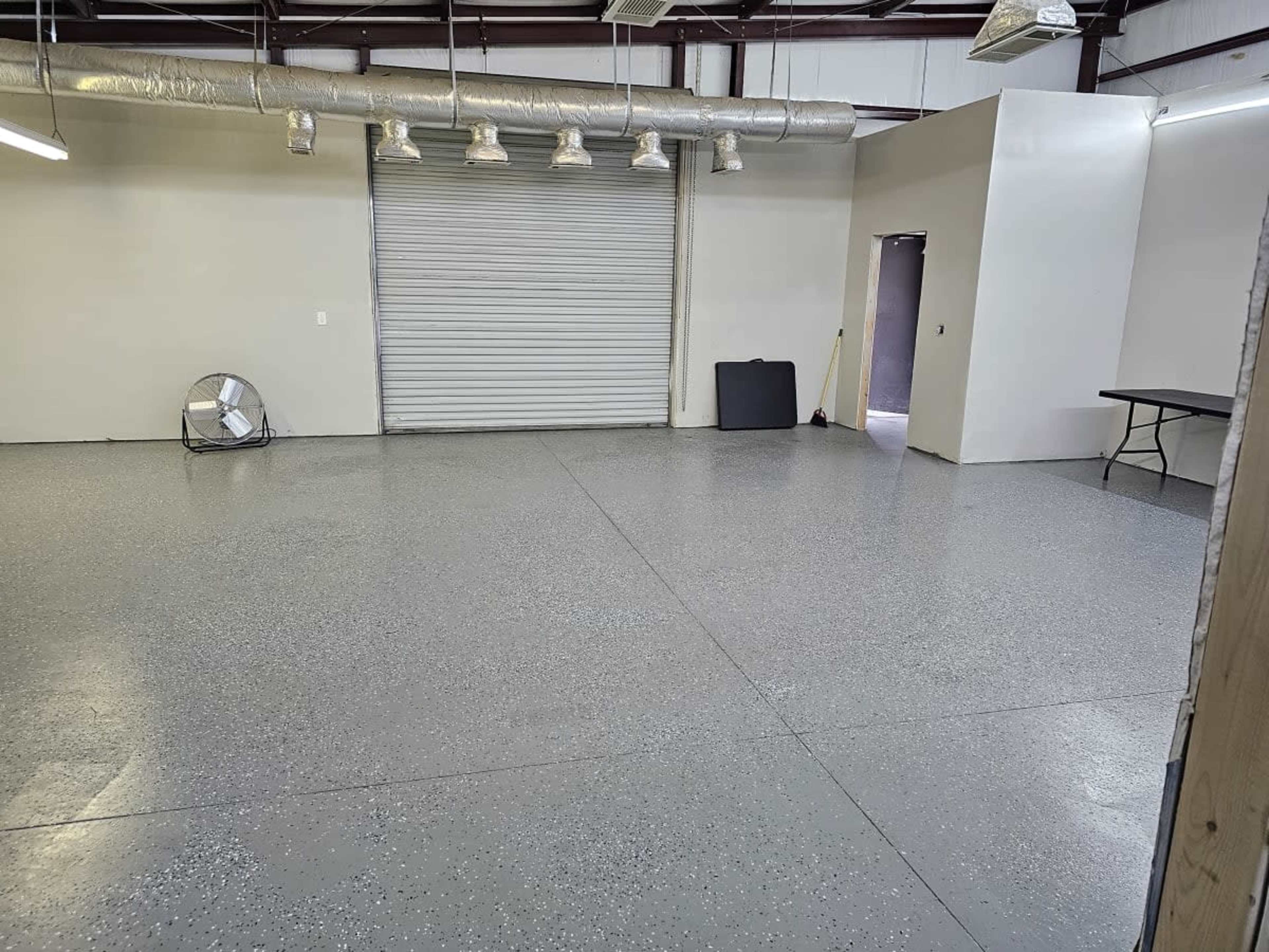 A large, empty industrial room with a gray floor, a closed roll-up door, and minimal furnishings.