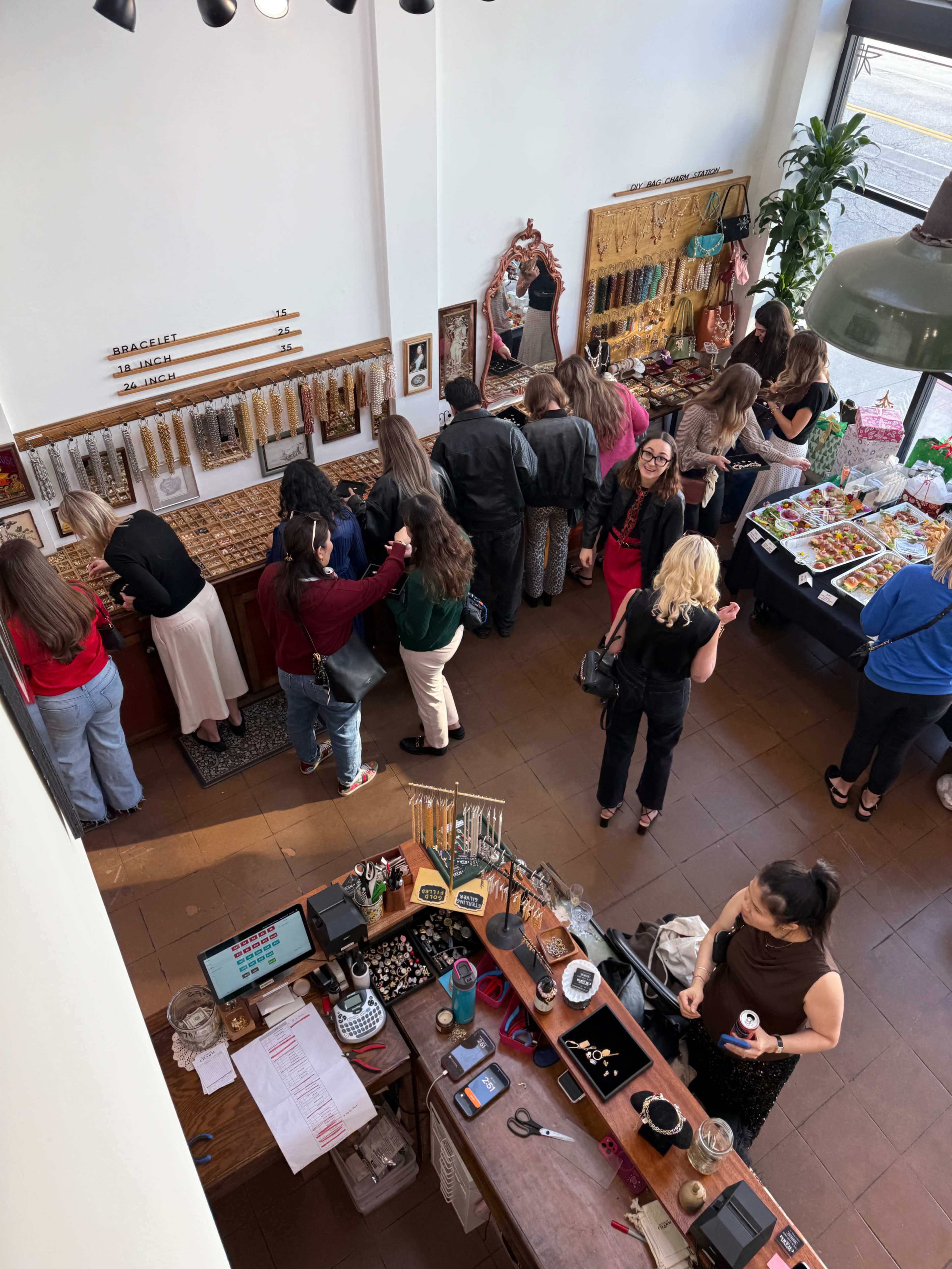 The image shows a busy boutique with a variety of people browsing jewelry displays and enjoying snacks at a table.