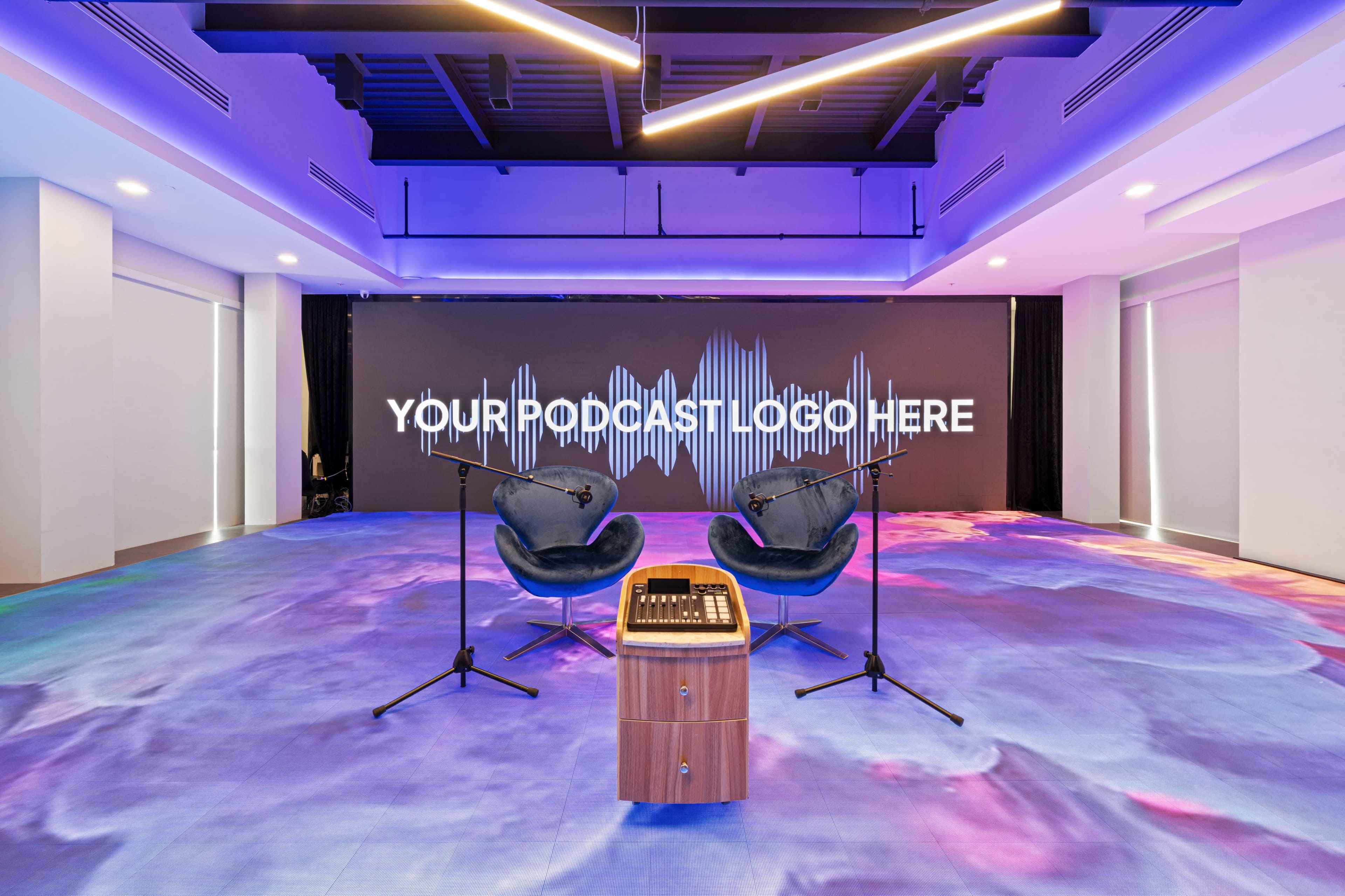The image features a modern podcast studio with two microphones on stands, two chairs facing a wall displaying a soundwave graphic and placeholder text for a podcast logo, all set against a colorful, artistic floor design.