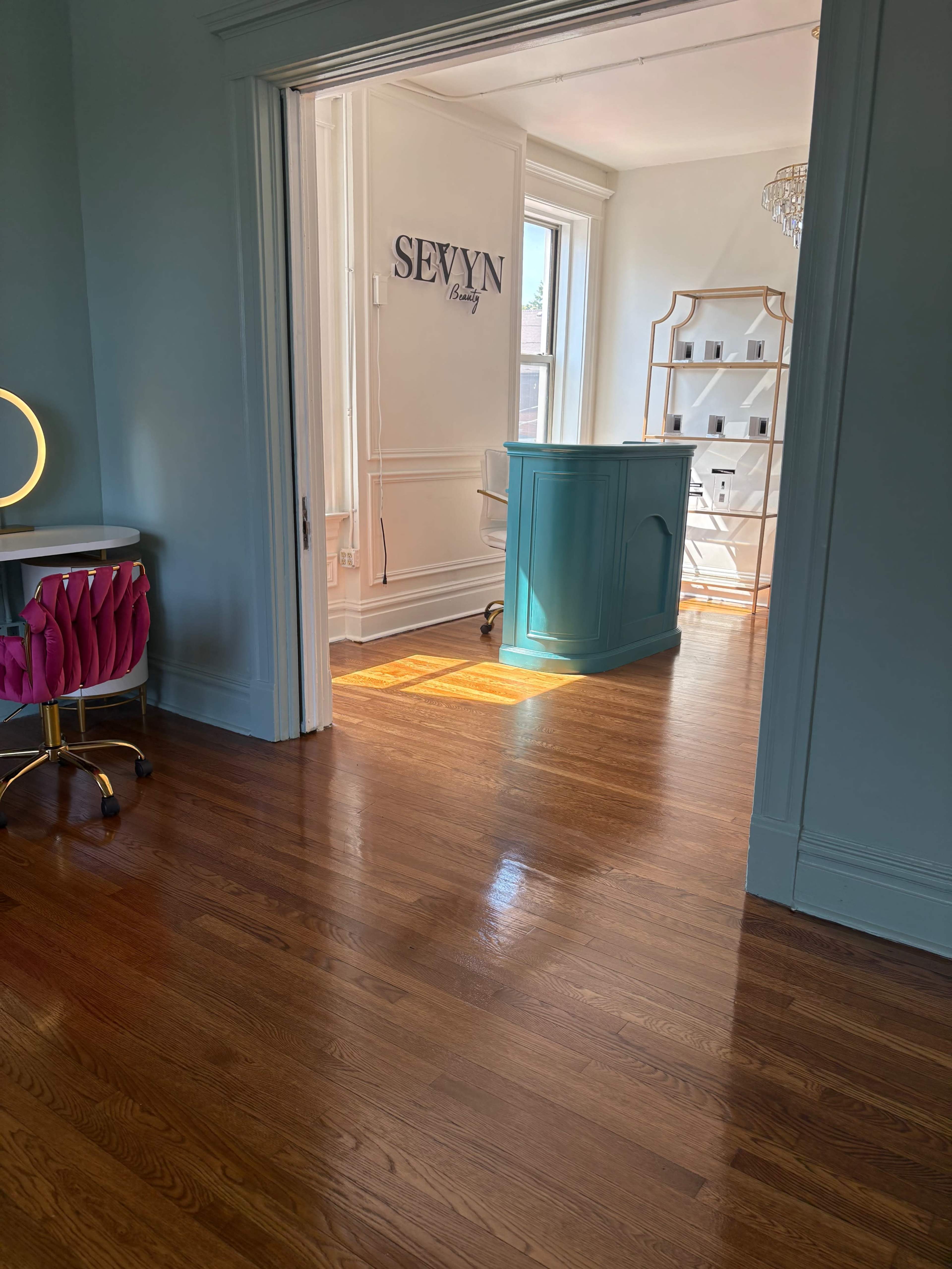 The image shows a light-filled room with a turquoise reception desk, a wooden floor, and a vanity station with a round mirror.