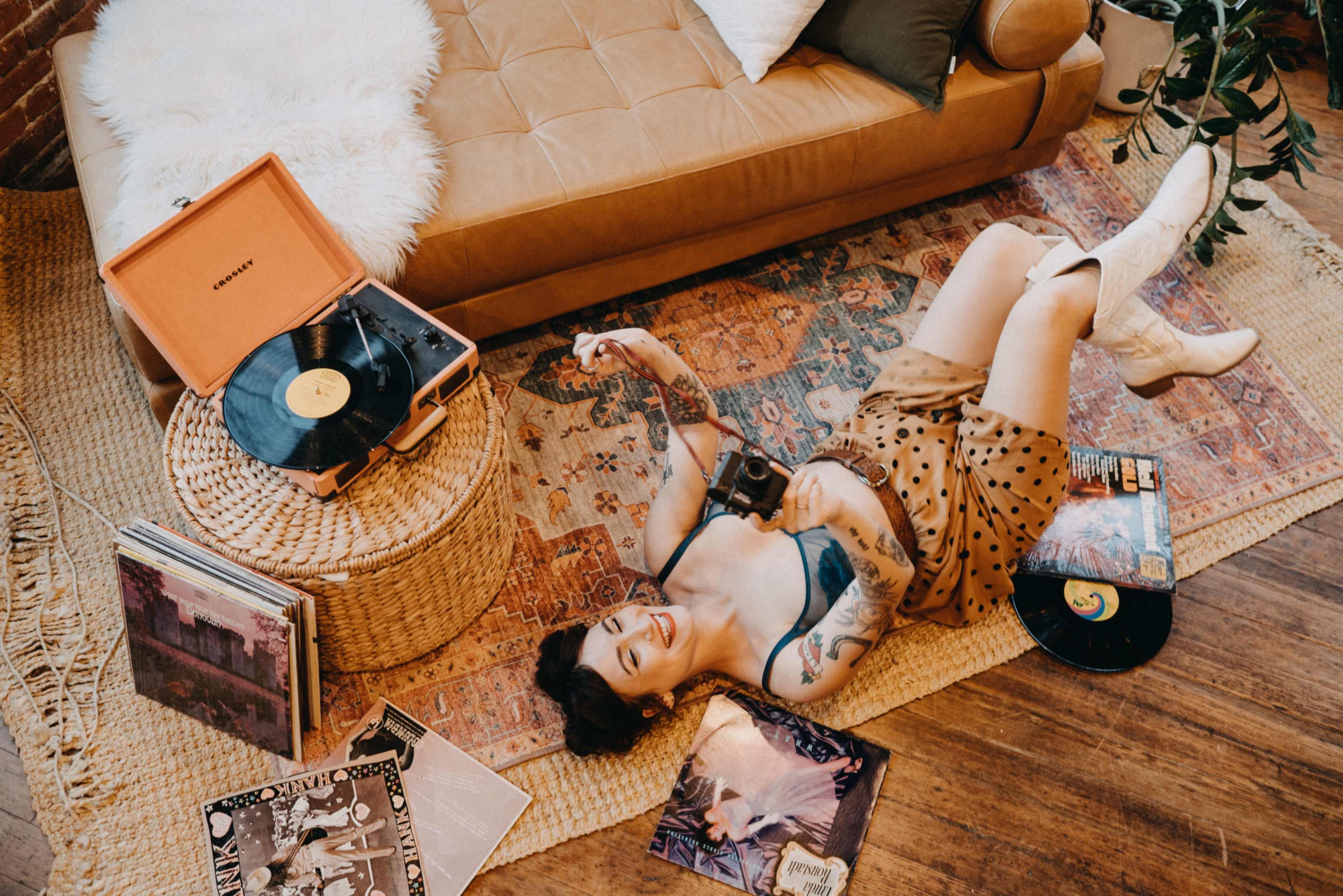 A woman with tattoos lies on a patterned rug surrounded by vinyl records and a retro record player in a cozy living room setting.