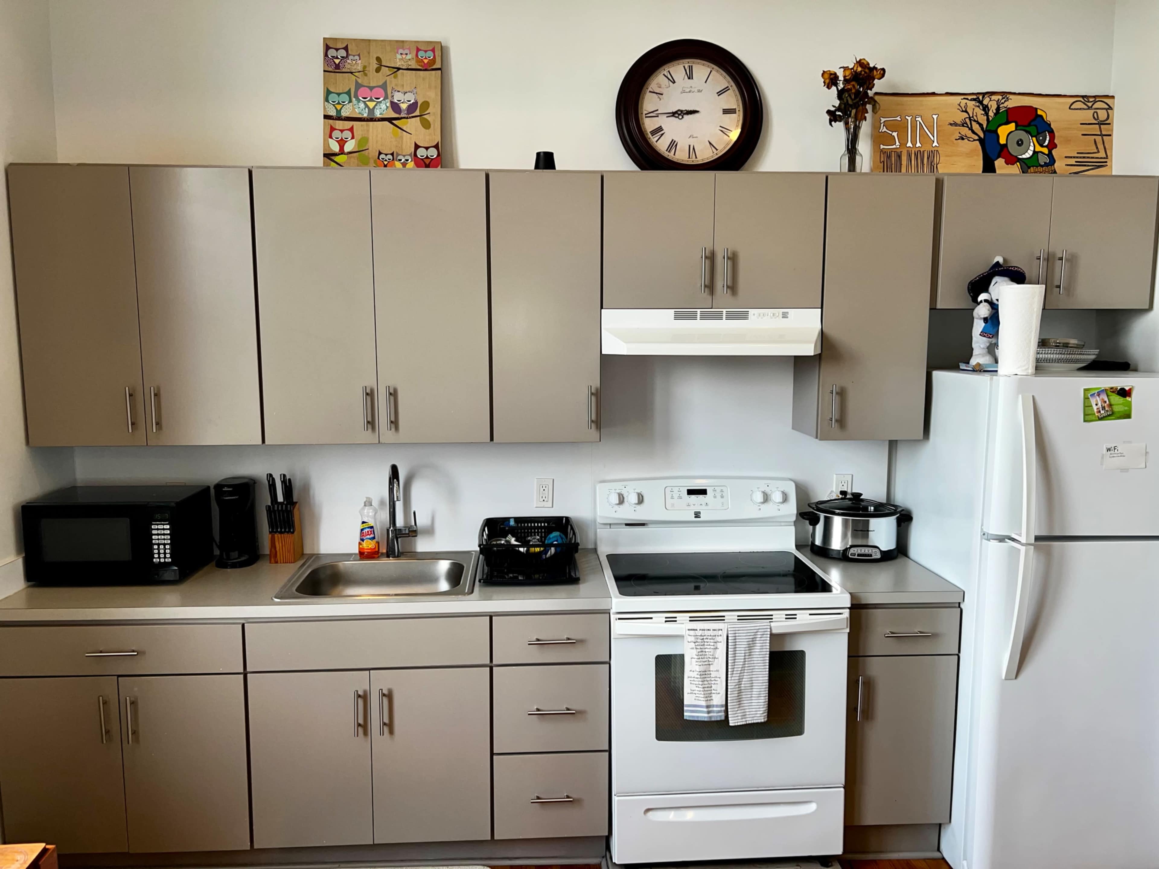 The image shows a modern kitchen with gray cabinetry, a stove, a microwave, and a refrigerator, along with a clock and decorative artwork on the walls.