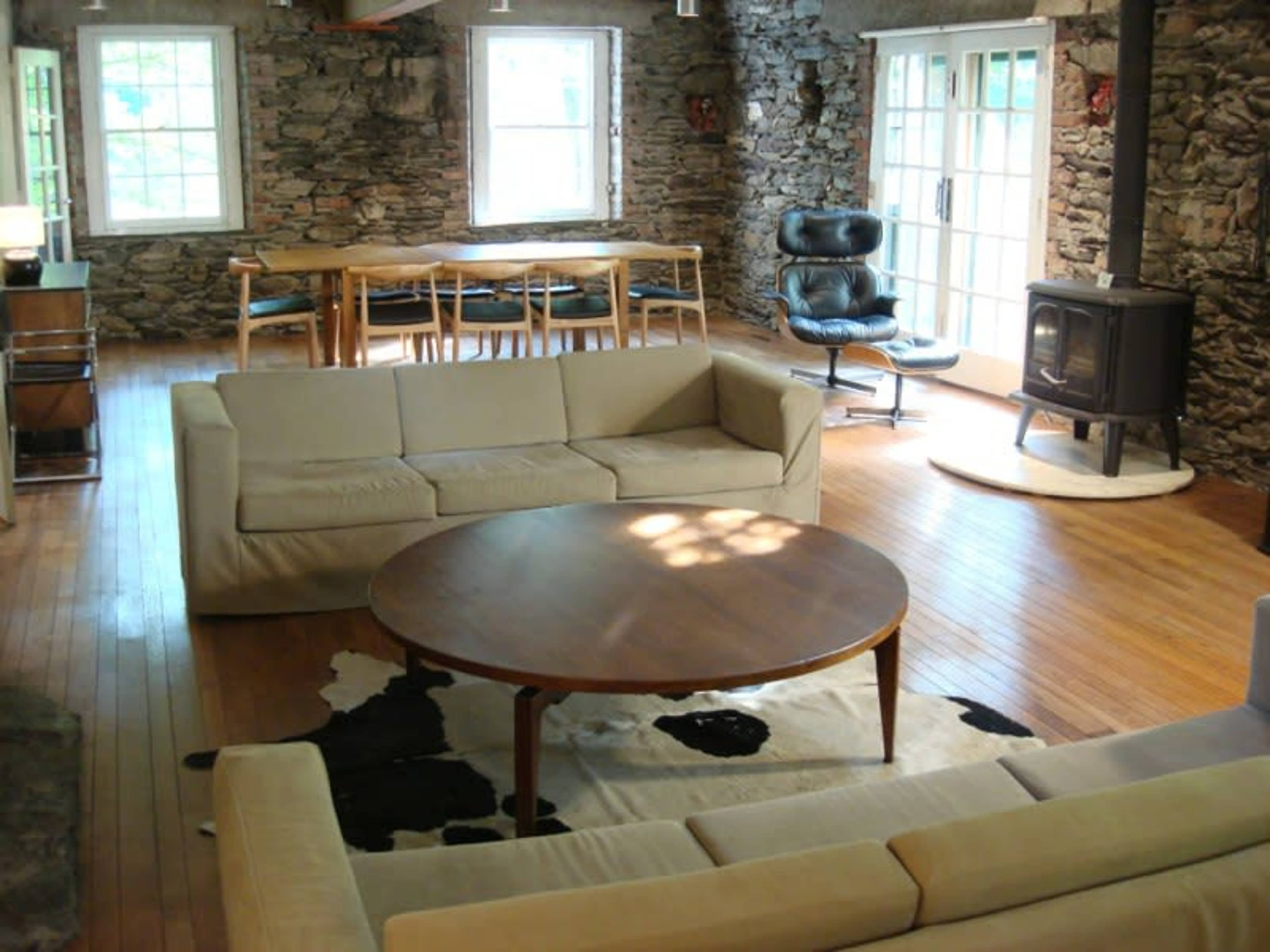The image shows a spacious living room featuring stone walls, wooden floors, a circular table, and a cozy seating area with a sofa and an armchair near a wood stove.