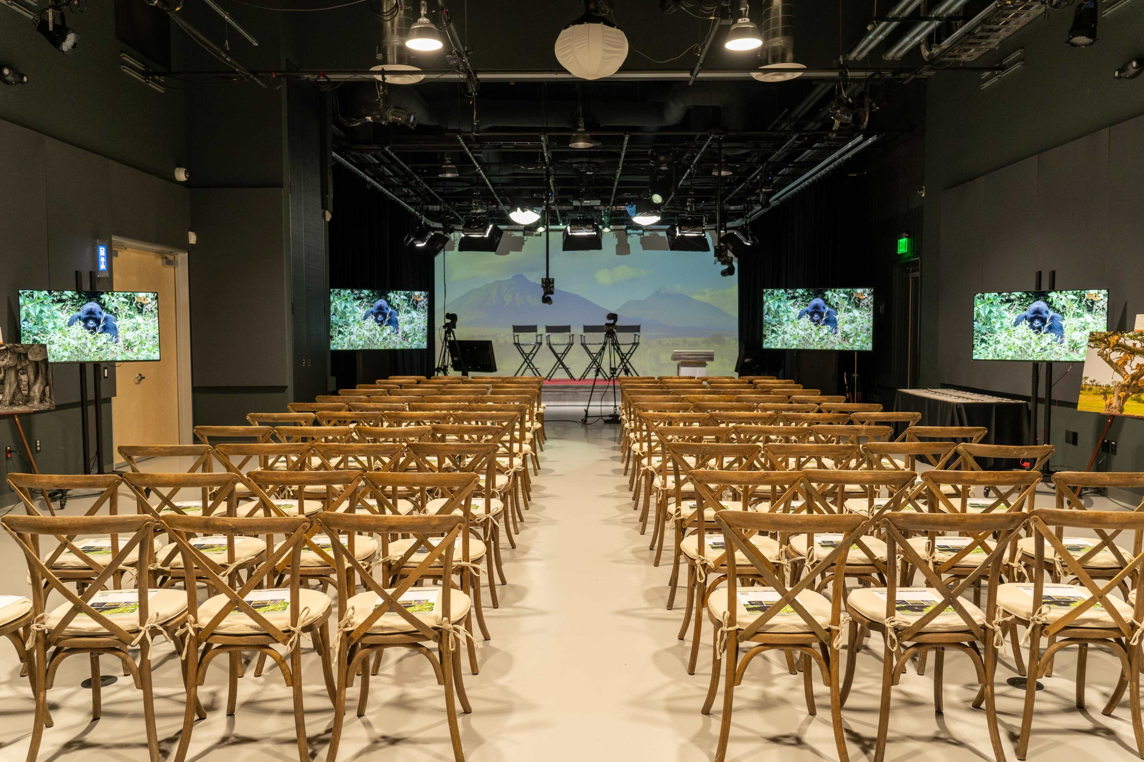 A conference room set up with rows of wooden chairs facing a stage, flanked by screens displaying nature imagery.