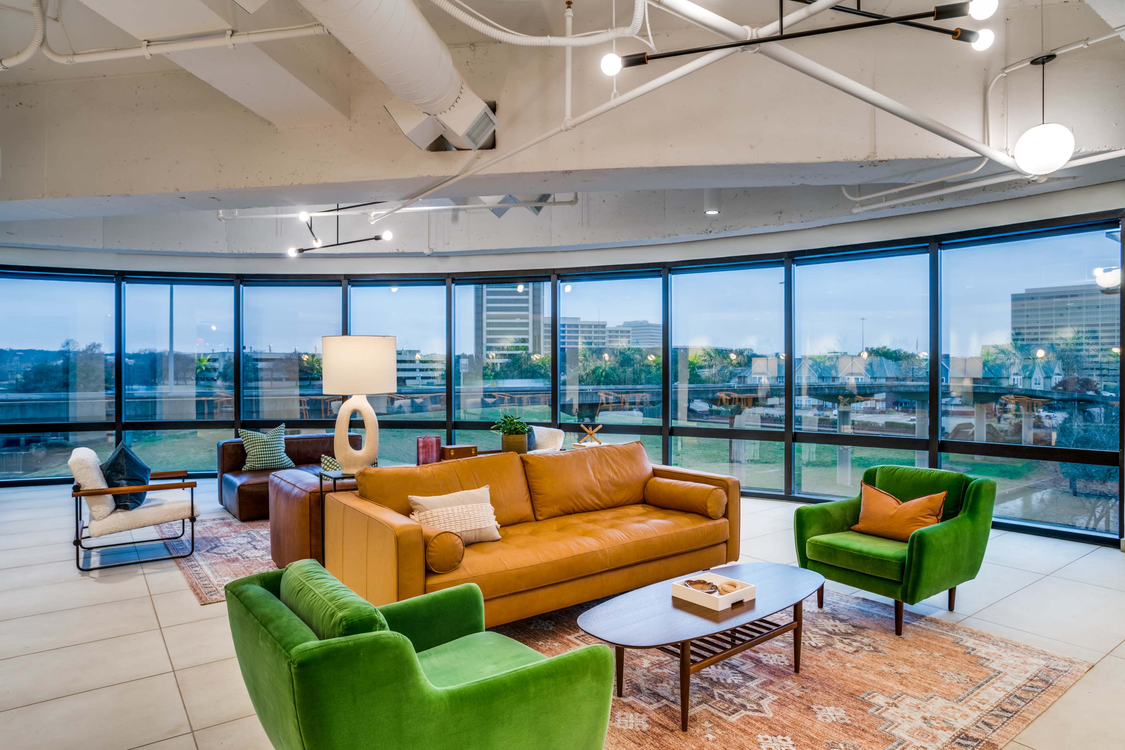 The image shows a modern, open lounge area with large windows, featuring a yellow sofa, green armchairs, a coffee table, and a distinctive lamp, set against an urban backdrop.