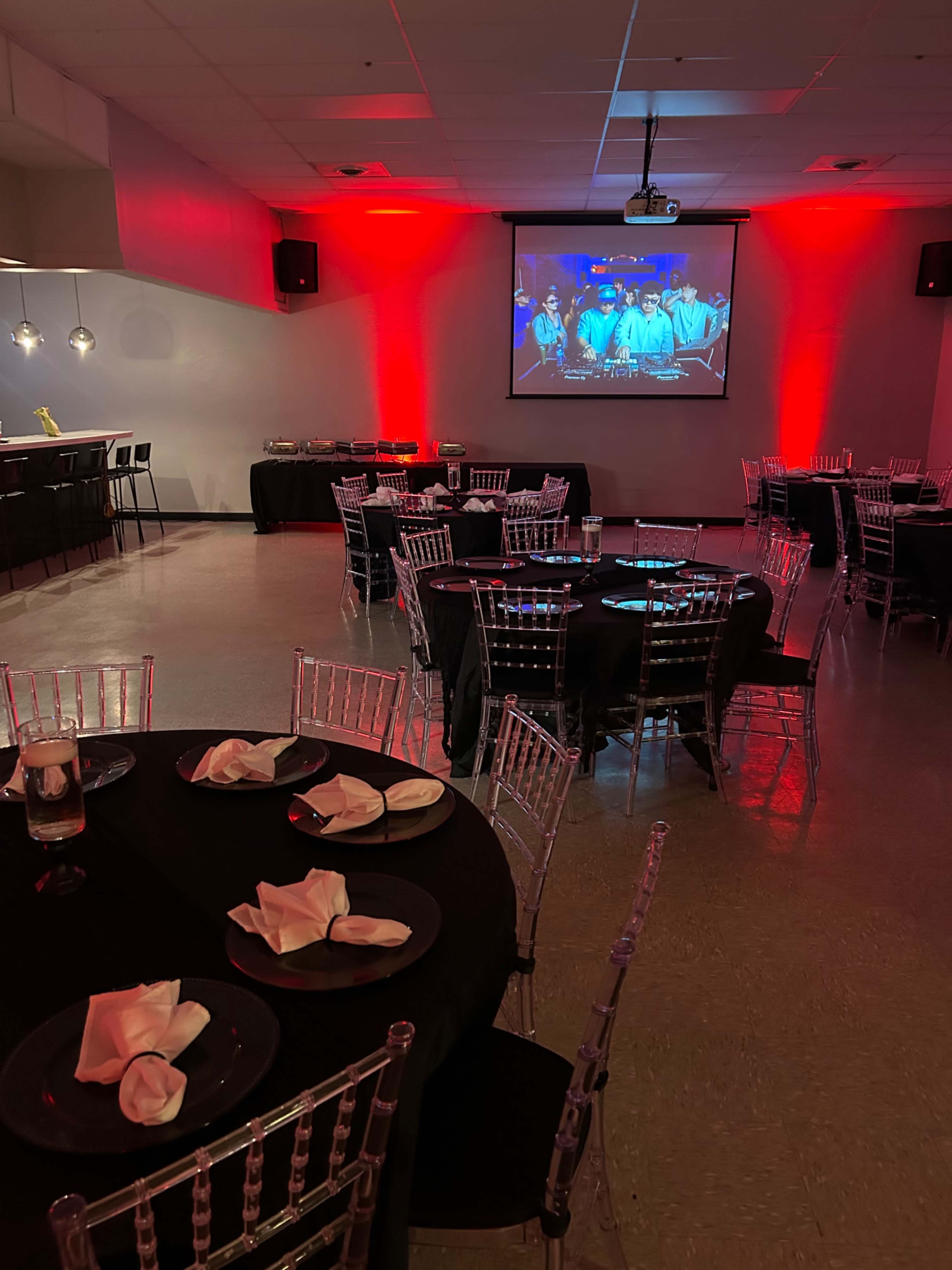 A banquet hall is set up with elegant, round tables covered in black cloth and surrounded by clear chiavari chairs, with a large screen displaying a presentation in the background.