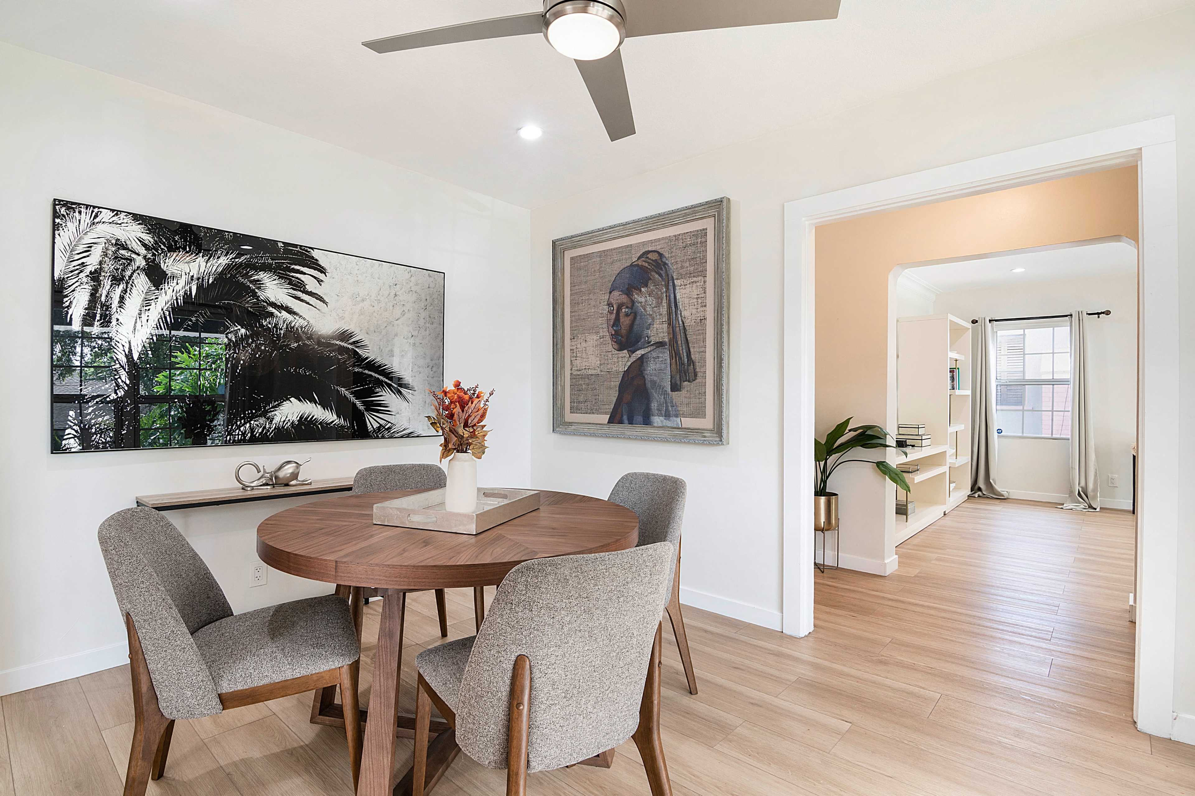 A dining area features a round wooden table surrounded by four upholstered chairs, with artwork and a doorway leading to another room.