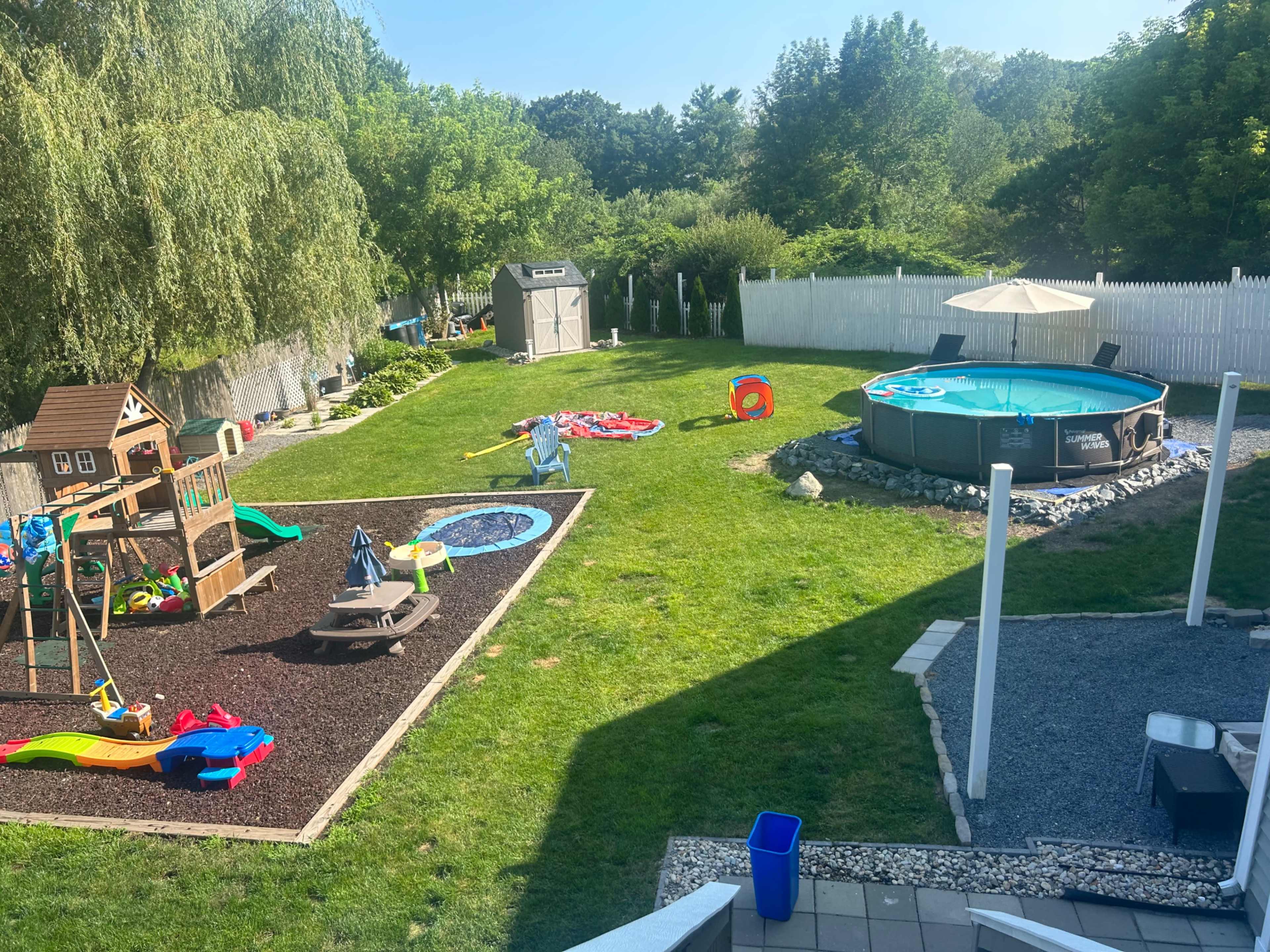 A backyard with a playset, a swimming pool, various toys scattered on the grass, and a shed in the background.