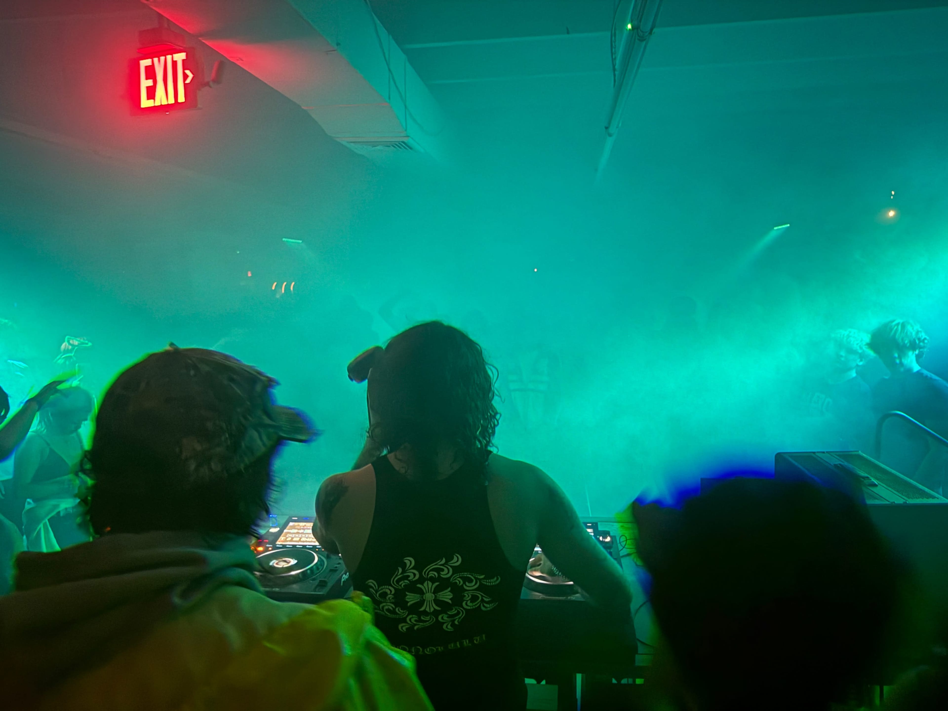 A DJ plays music in a dimly lit venue filled with fog and colorful lighting, while a crowd enjoys the performance.