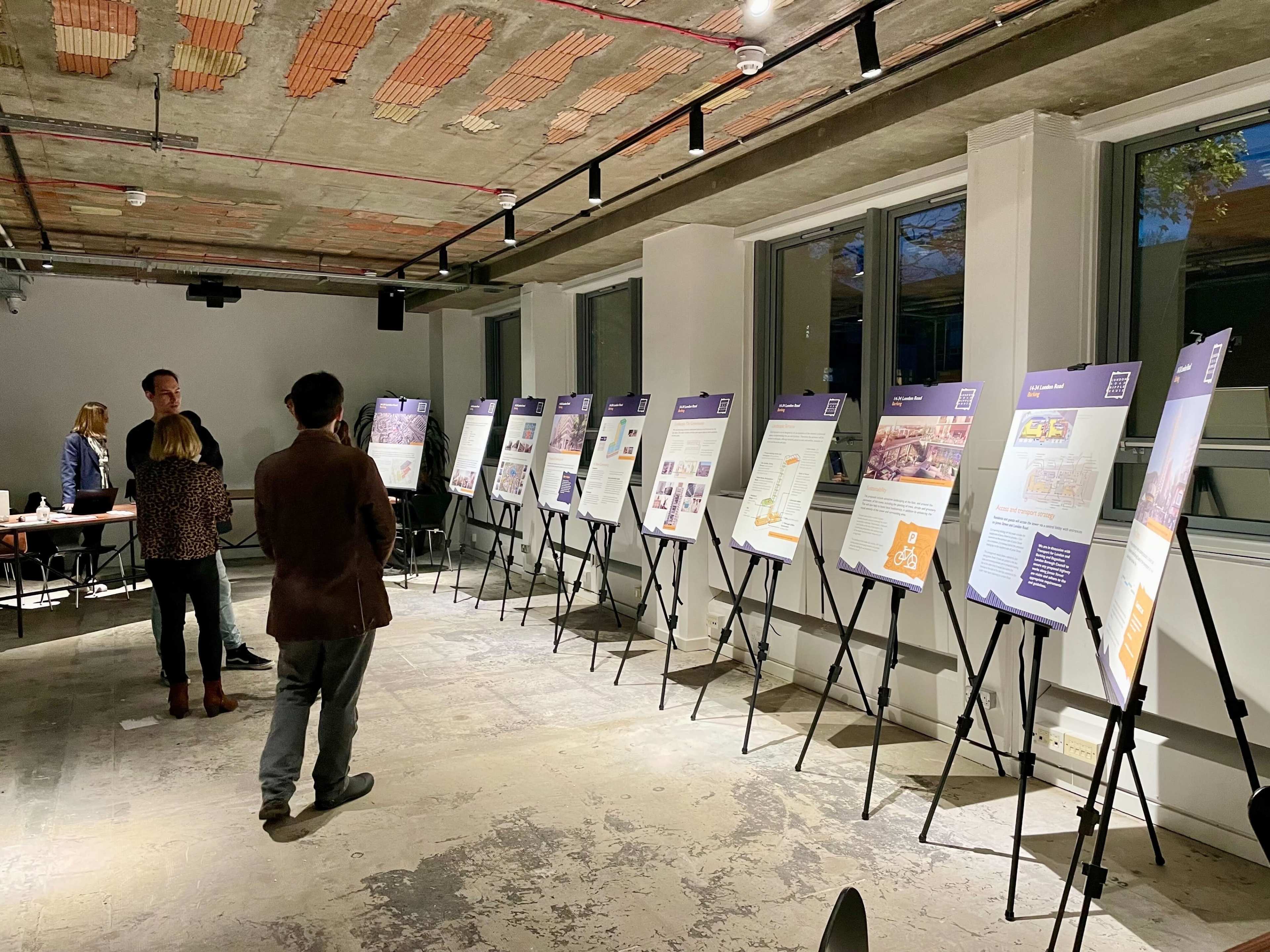 The image shows a dimly lit room with several informational display boards arranged in a row, while a few people are engaged in conversation near the boards.