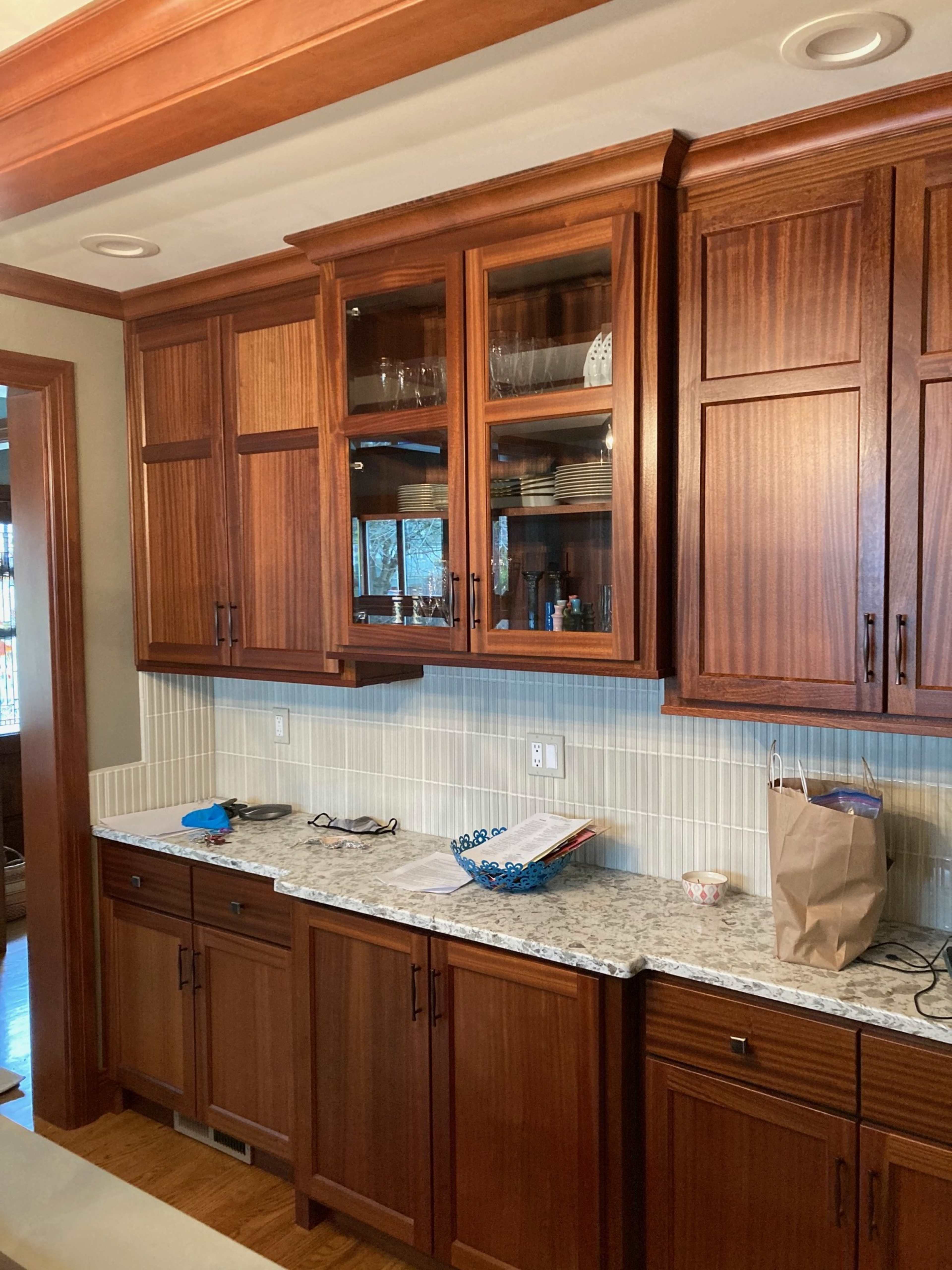 The image shows a kitchen with wooden cabinets, a granite countertop, and a tiled backsplash, featuring glassware and dishes displayed in an upper cabinet.
