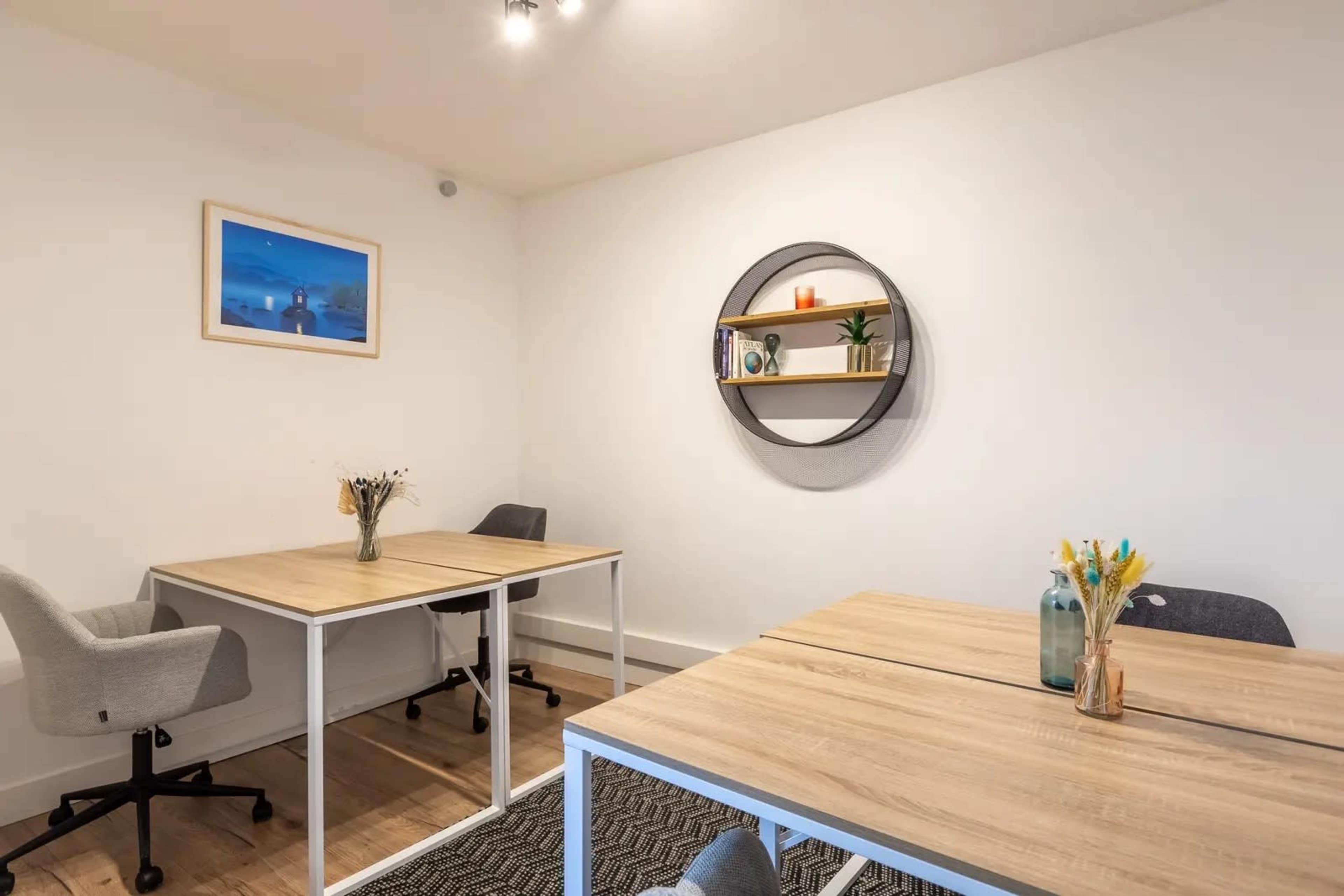 The image shows a minimalistic office space with two wooden tables, each adorned with a vase of flowers, and a circular shelf on the wall displaying small plants and decor.