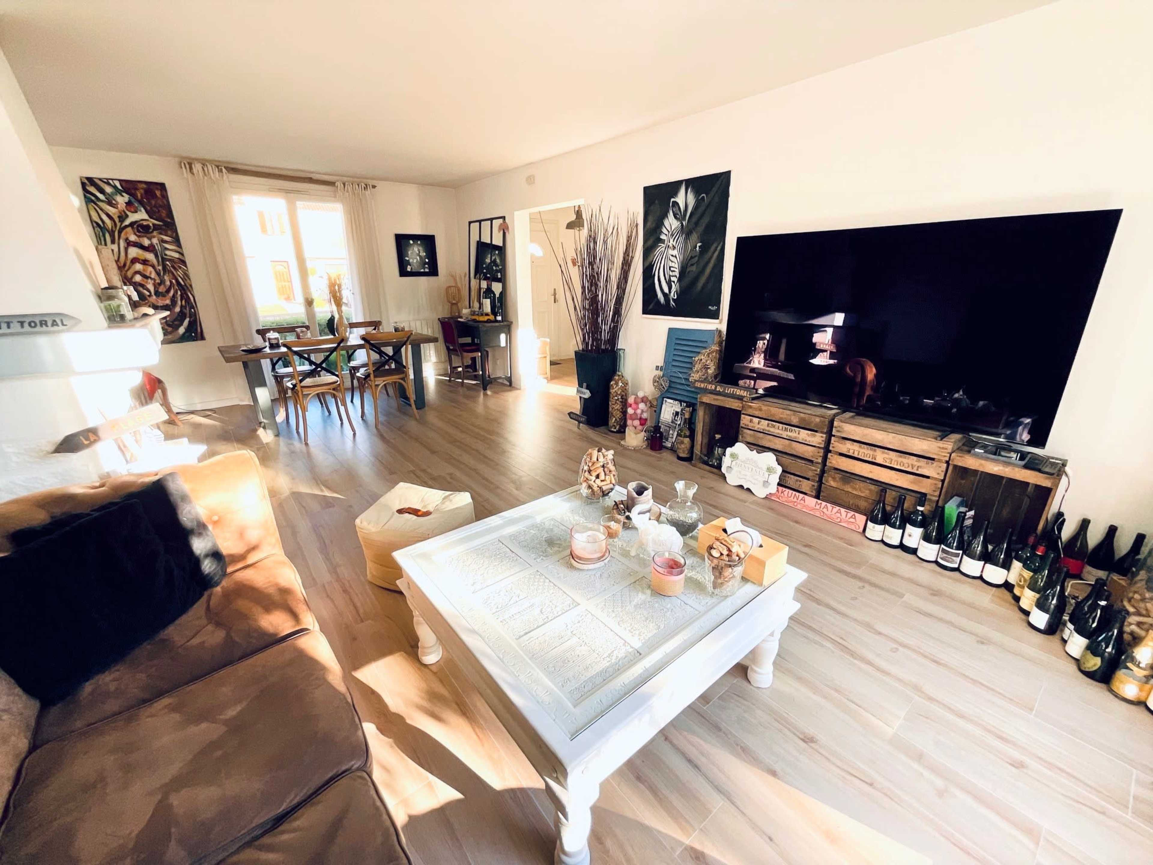 The living room features a large TV on a wooden stand, a white coffee table, a brown sofa, and a dining area with a table surrounded by chairs, illuminated by natural light from a window.