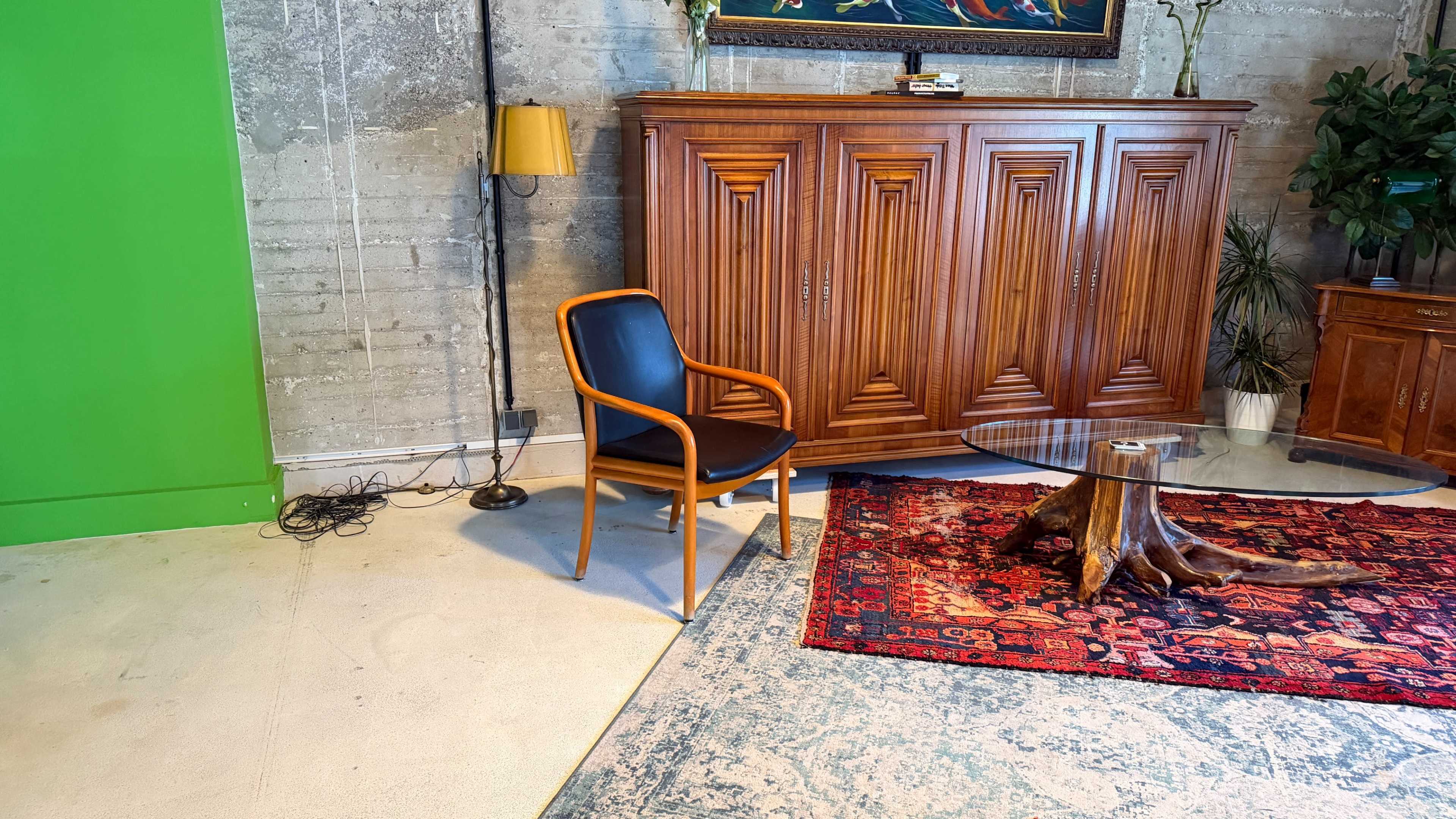 The image shows a modern interior space featuring a wooden cabinet, a black chair, a glass coffee table on a rustic wooden base, and a colorful rug on the floor.