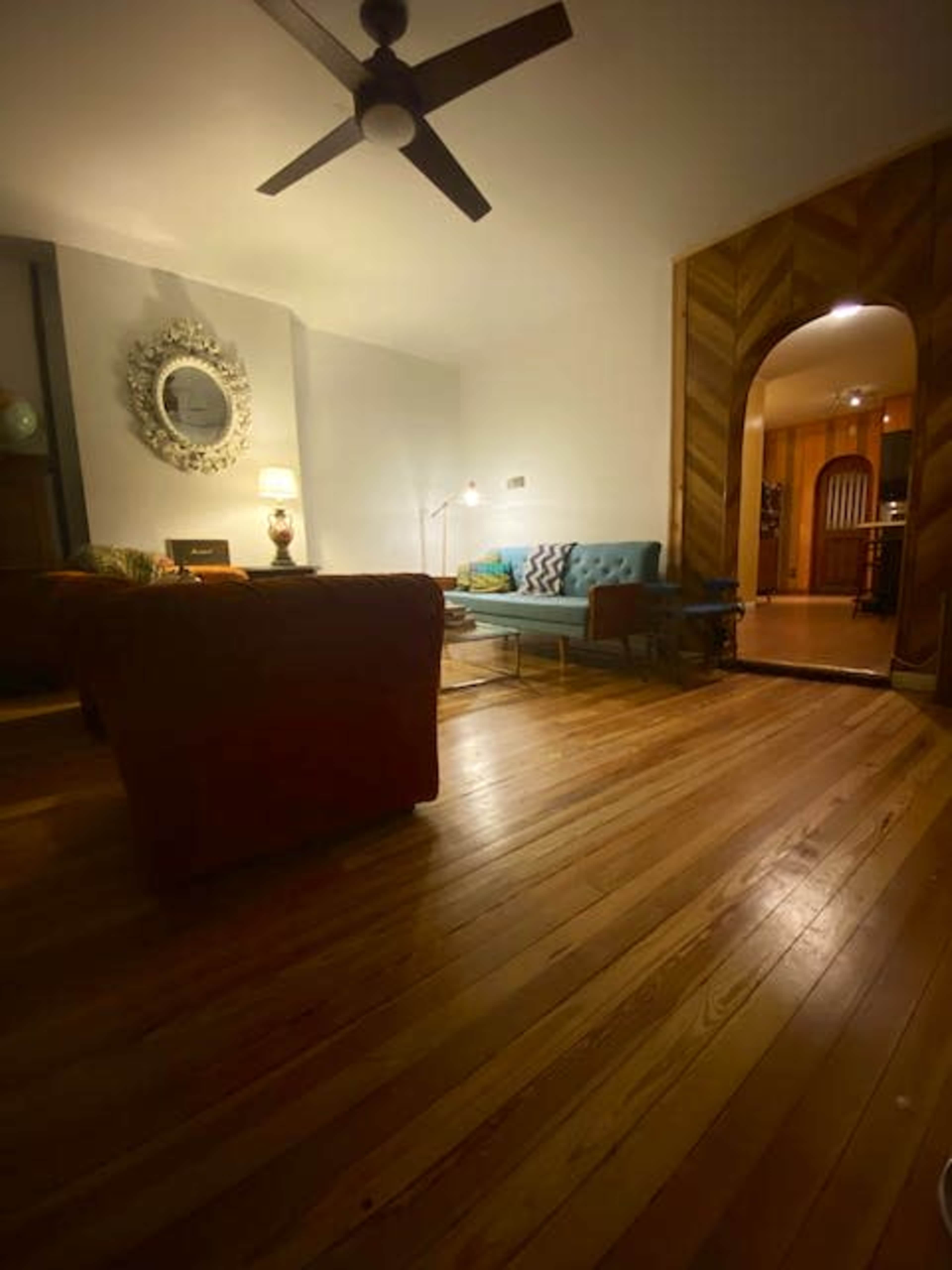 A living room with wooden floors, a ceiling fan, a large mirror on the wall, a blue couch, and an arched entryway leading to another room.