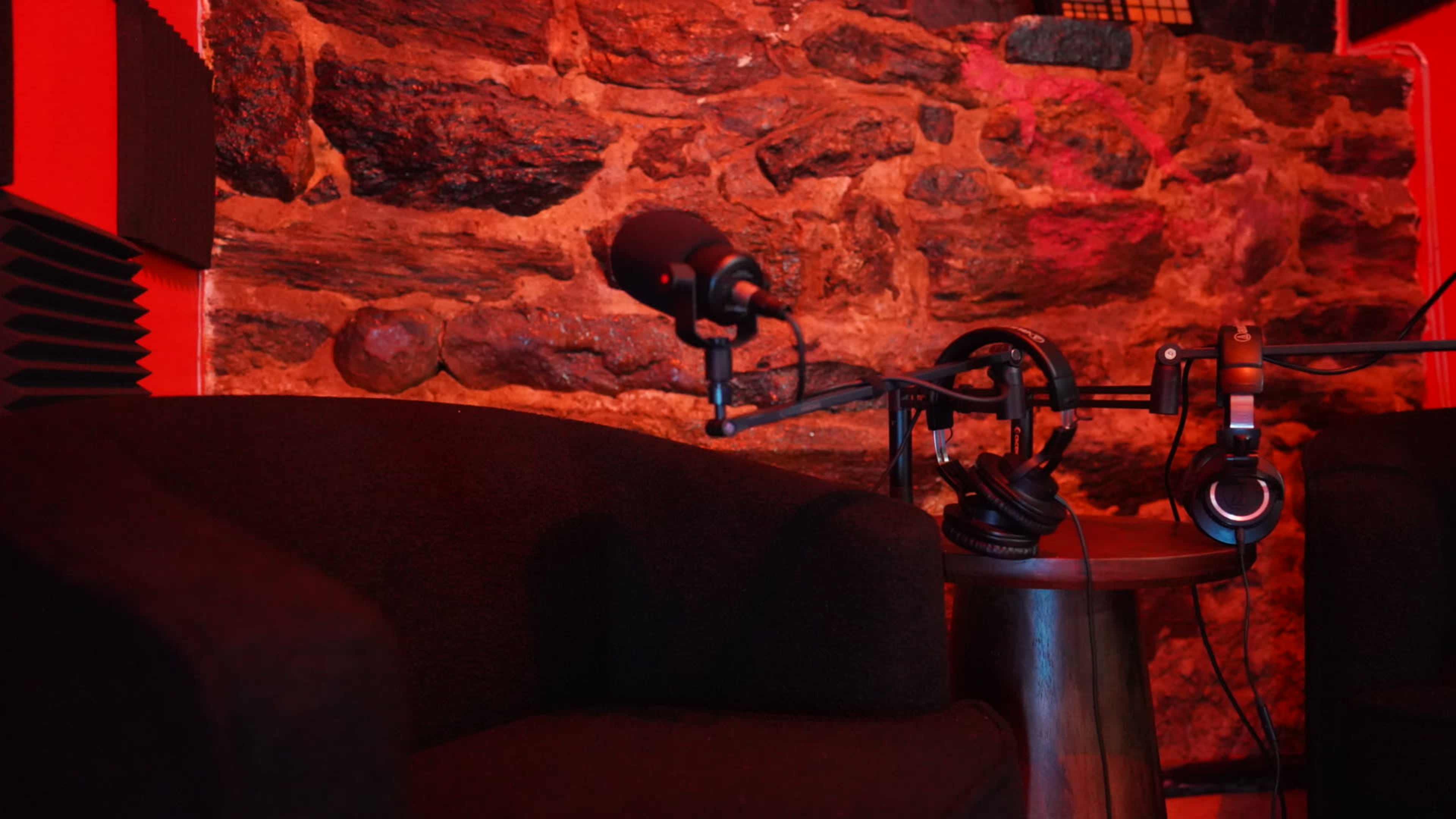 The image shows a dimly lit recording studio with a black chair, a microphone on a stand, and a pair of headphones resting on a table beside a stone wall.