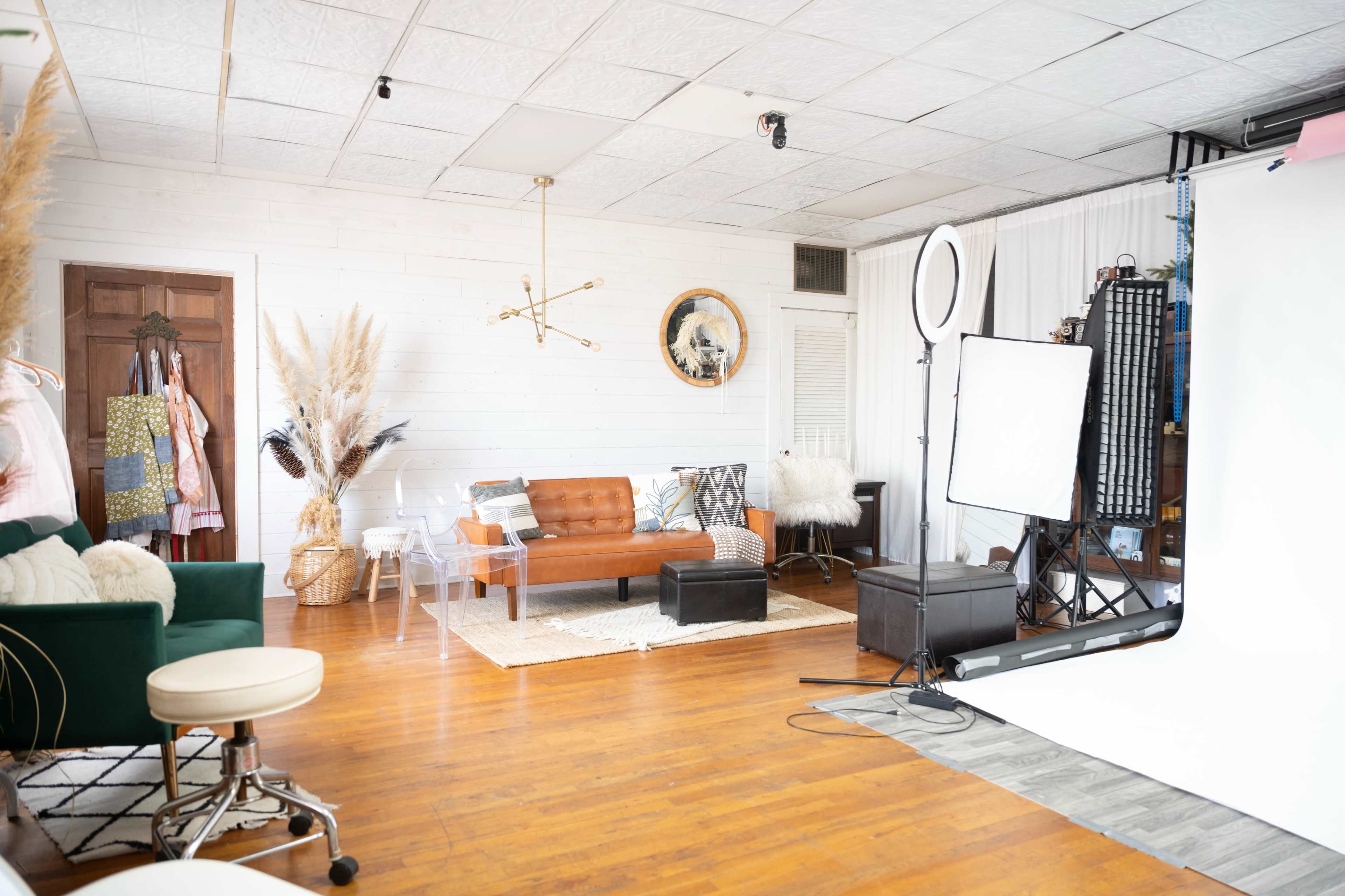 The image shows a well-lit photography studio featuring a brown sofa, decorative plants, a light setup, and a variety of clothing items hanging on a rack.