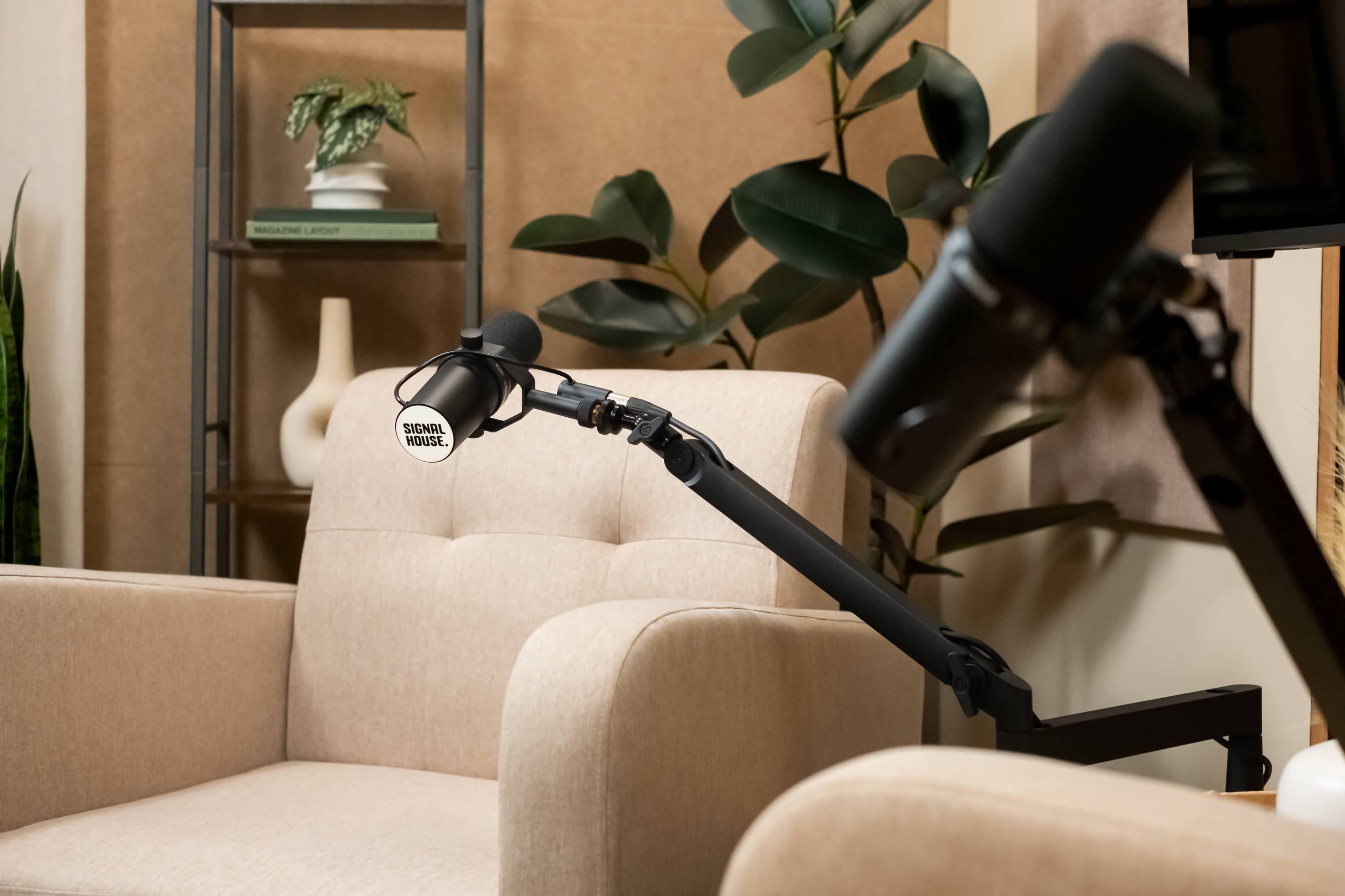 The image shows a cozy, minimalistic recording studio setup featuring a beige armchair, a microphone on an adjustable stand, and greenery in the background.