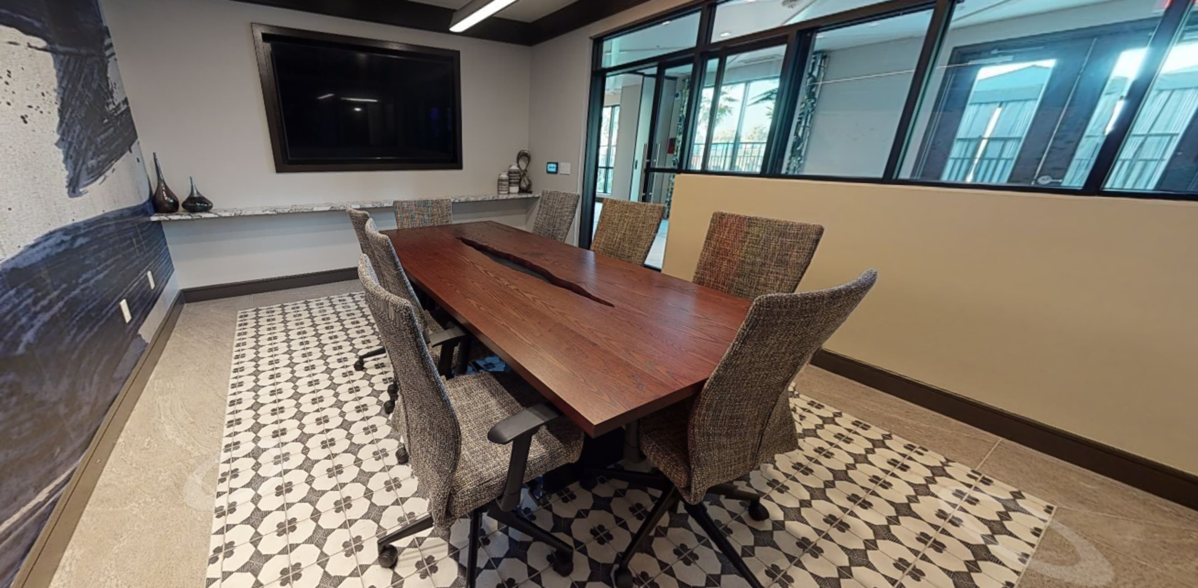 A conference room features a long wooden table surrounded by six chairs, with large windows and a patterned floor.