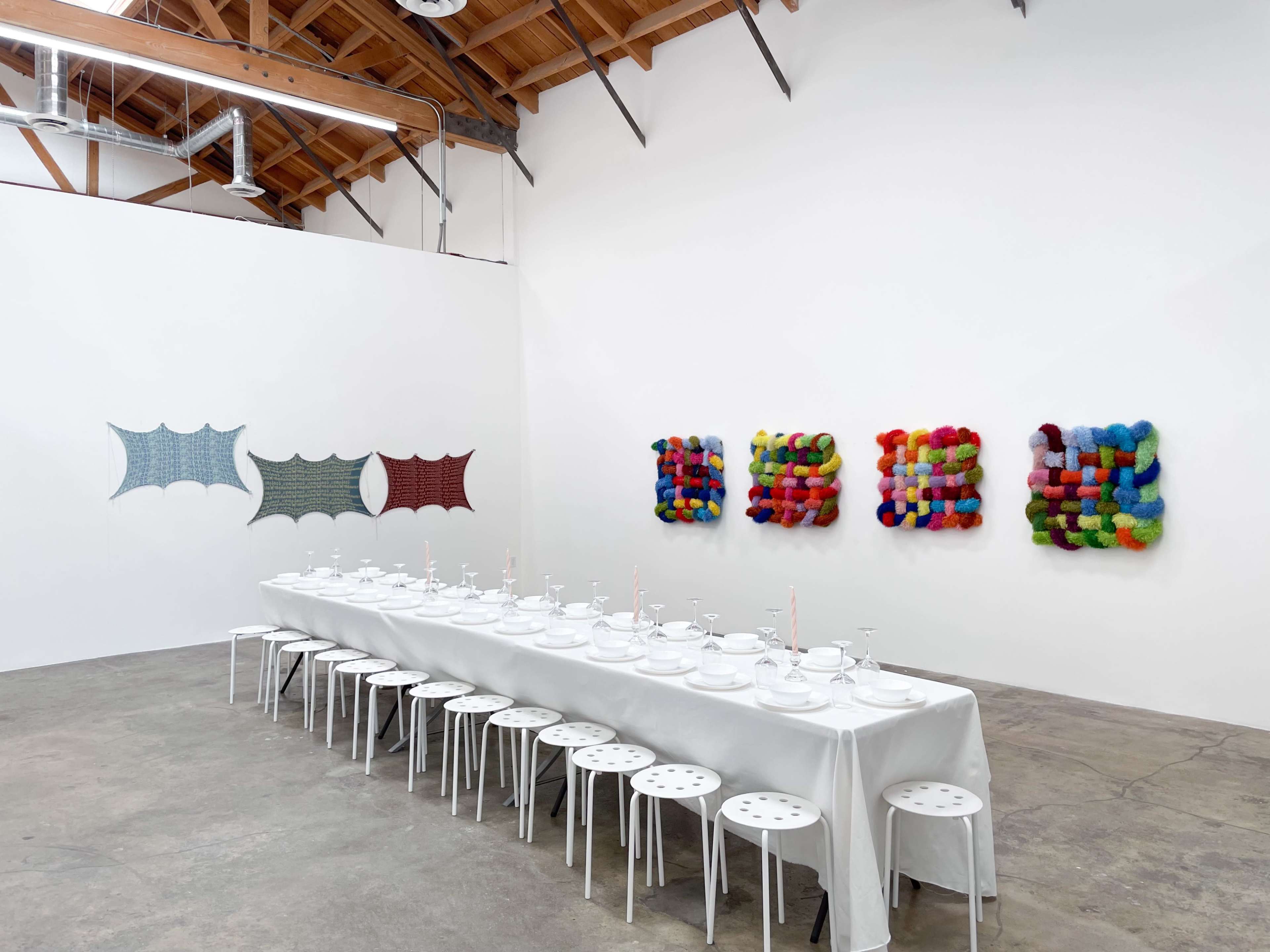 A long, white dining table set with plates and glasses occupies the center of an art gallery, surrounded by colorful textile artwork on the walls.