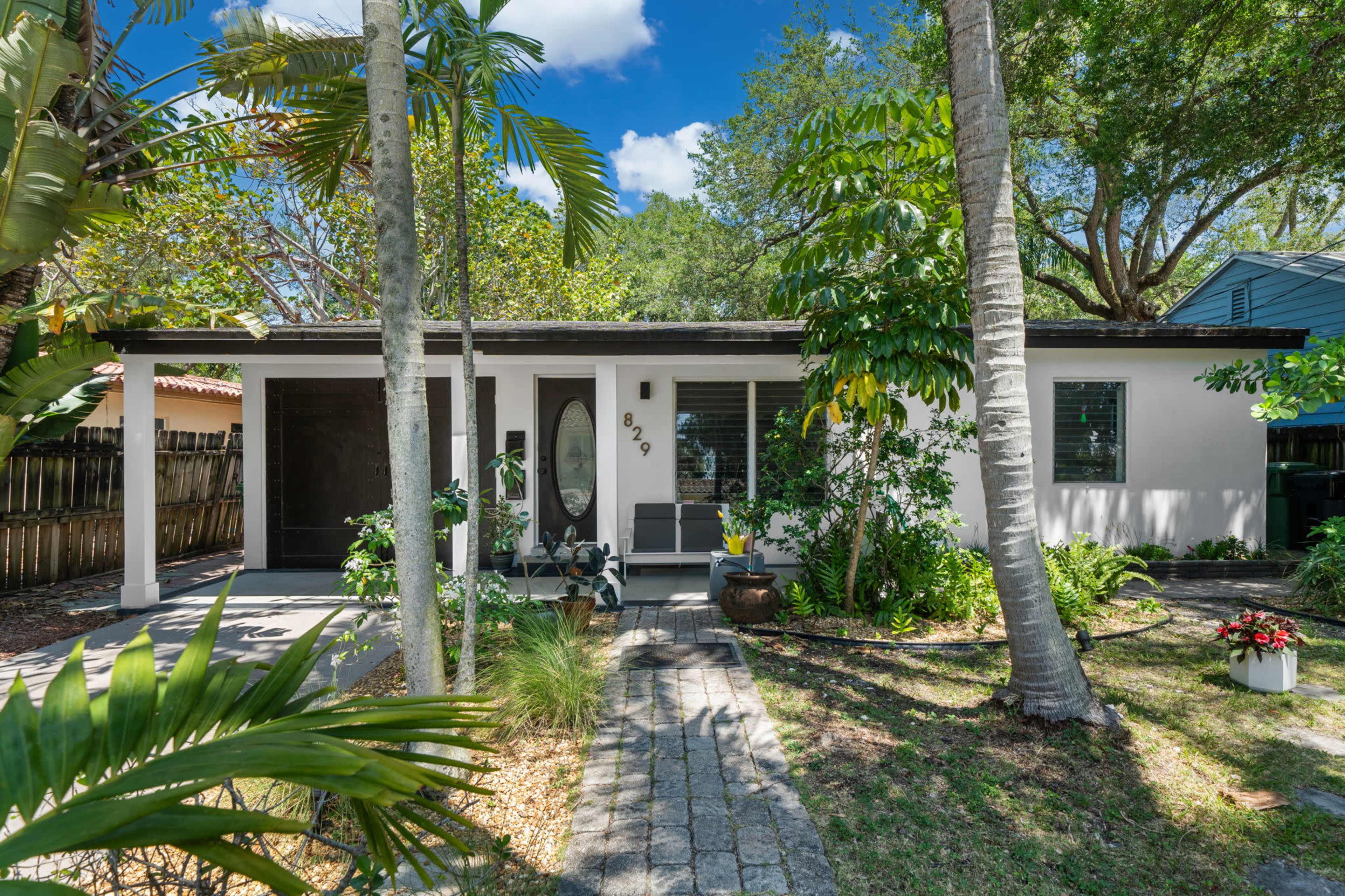 A single-story house with a front porch, surrounded by greenery and palm trees.