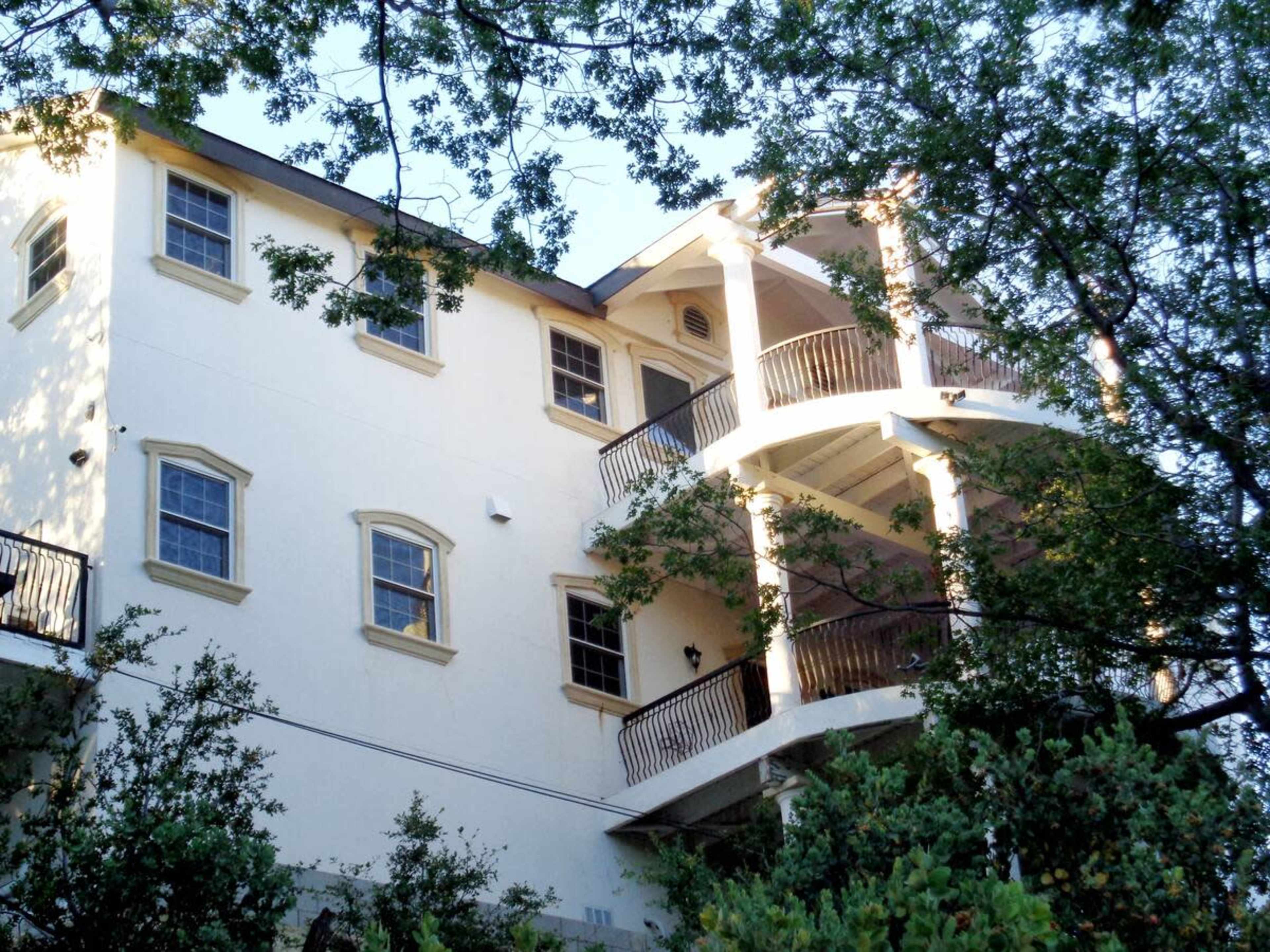 The image shows a three-story house with a white exterior featuring multiple balconies and surrounded by trees.