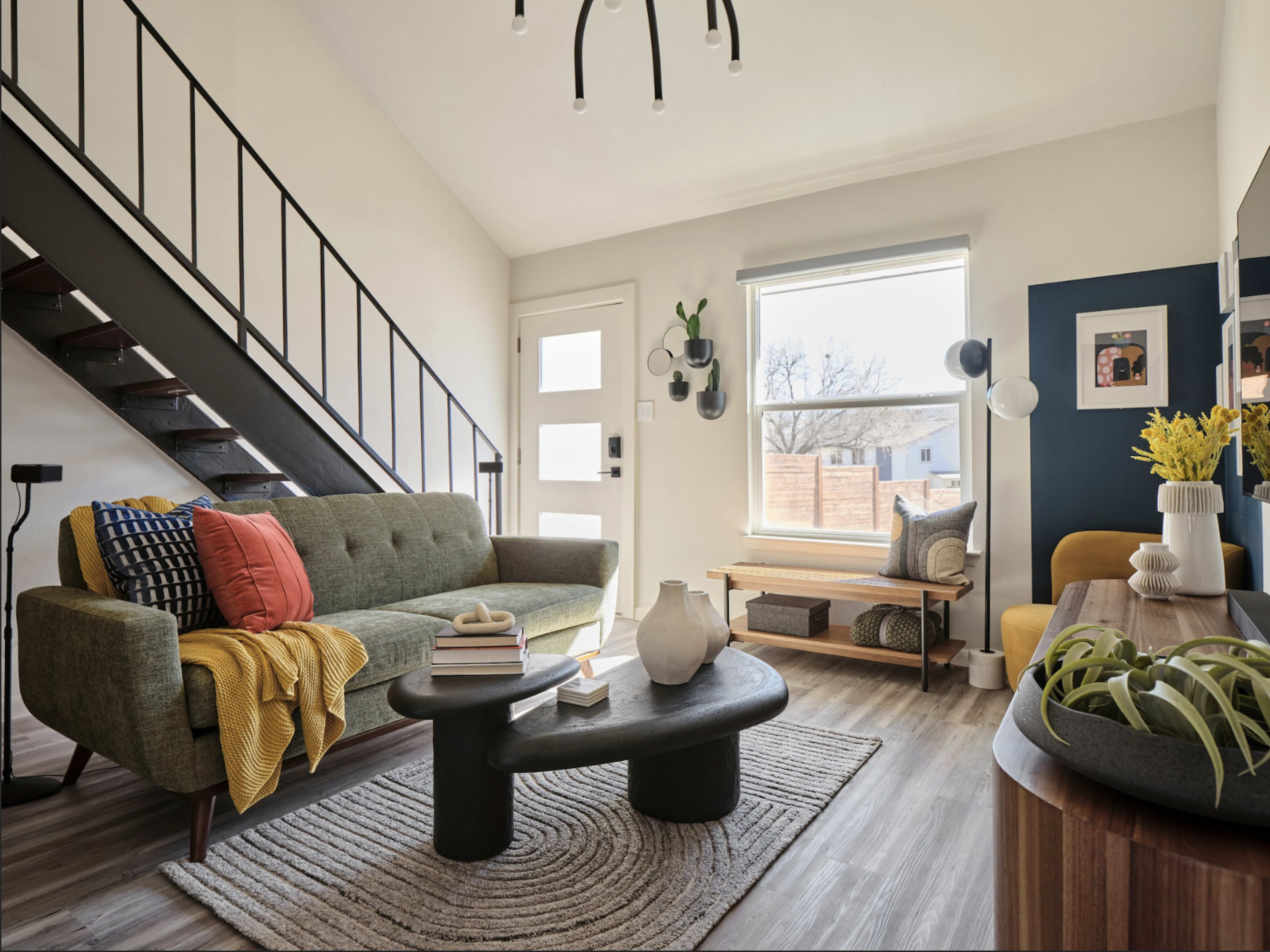 The living room features a green sofa, a round black coffee table, and a staircase, with large windows letting in natural light.