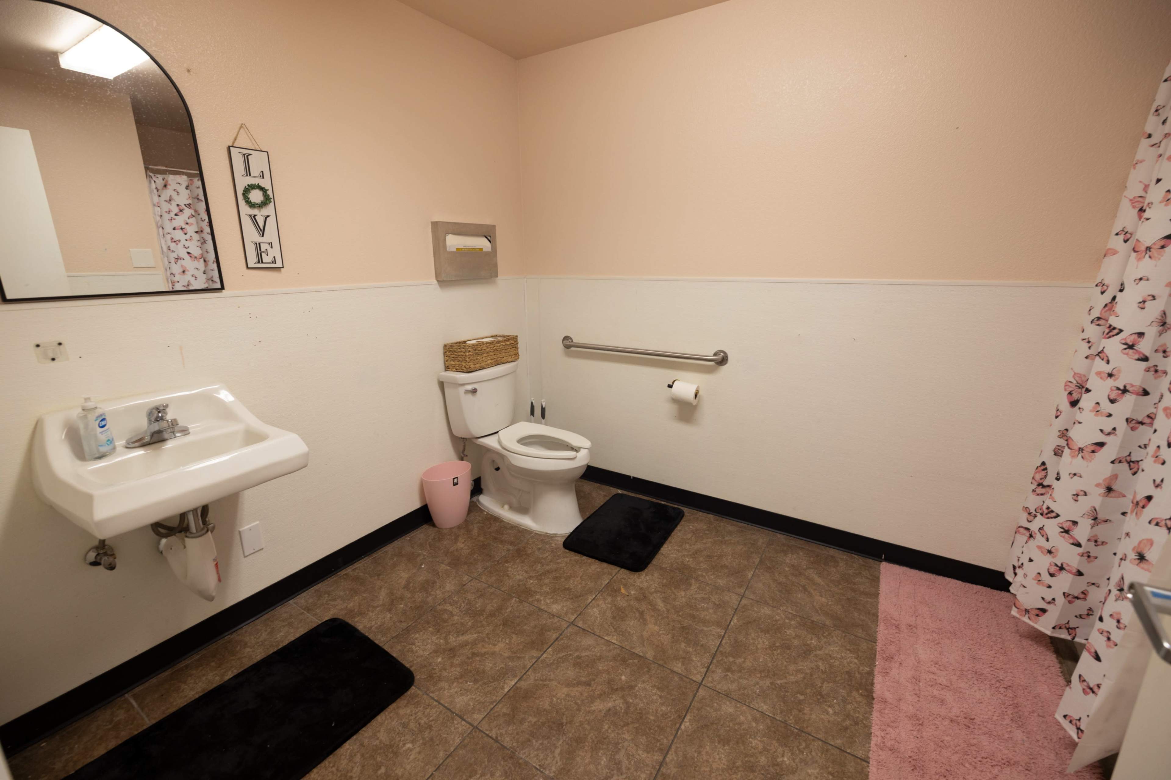 The image shows a clean bathroom featuring a sink, toilet, and a shower curtain with a floral pattern, all set against pink walls and tile flooring.