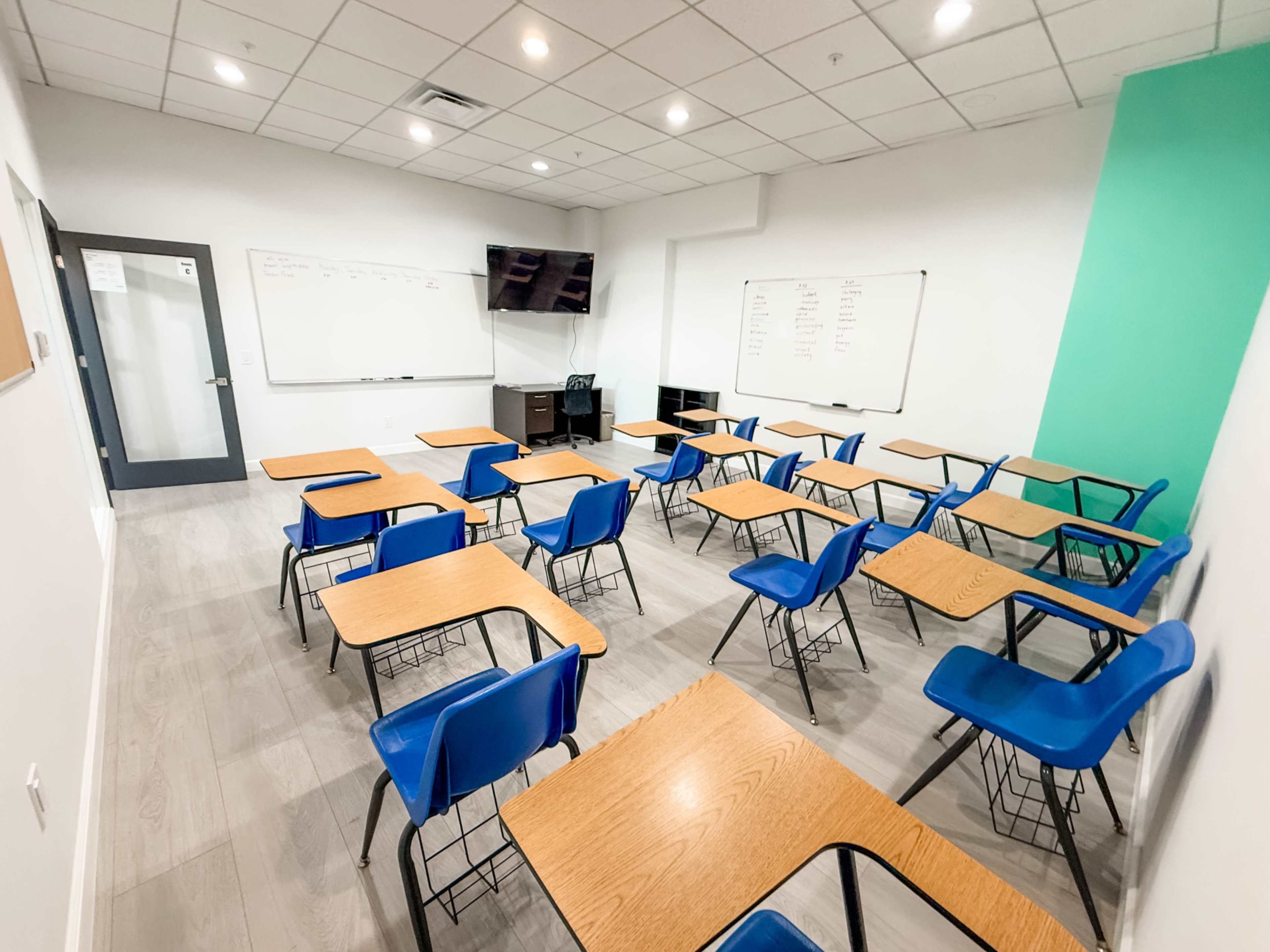 The image shows a classroom with rows of blue desks and chairs, a television mounted on the wall, and whiteboards on the front.