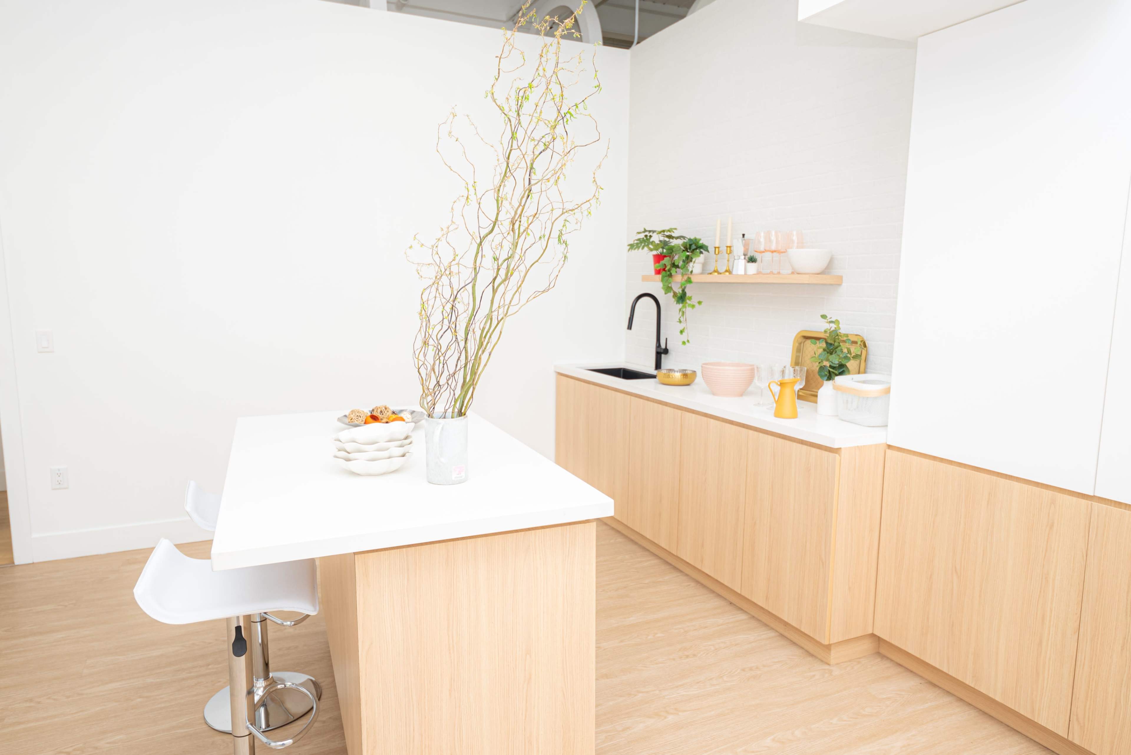 A bright kitchen features a white island with bar stools, light wood cabinetry, and decorative plants on the counters.