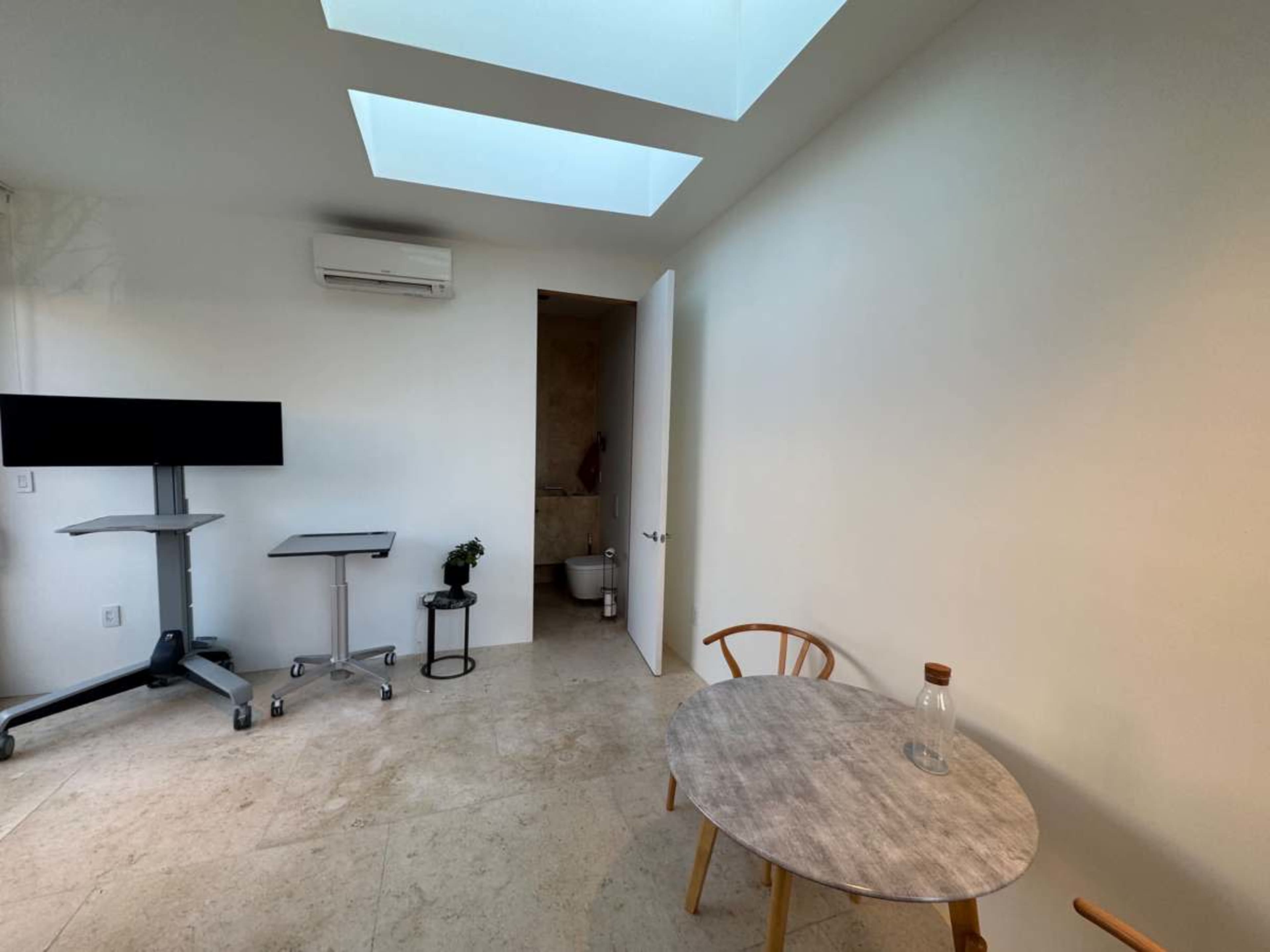 The image shows a minimalist room with a small round table and chairs, an air conditioning unit on the wall, and an open door leading to a bathroom.