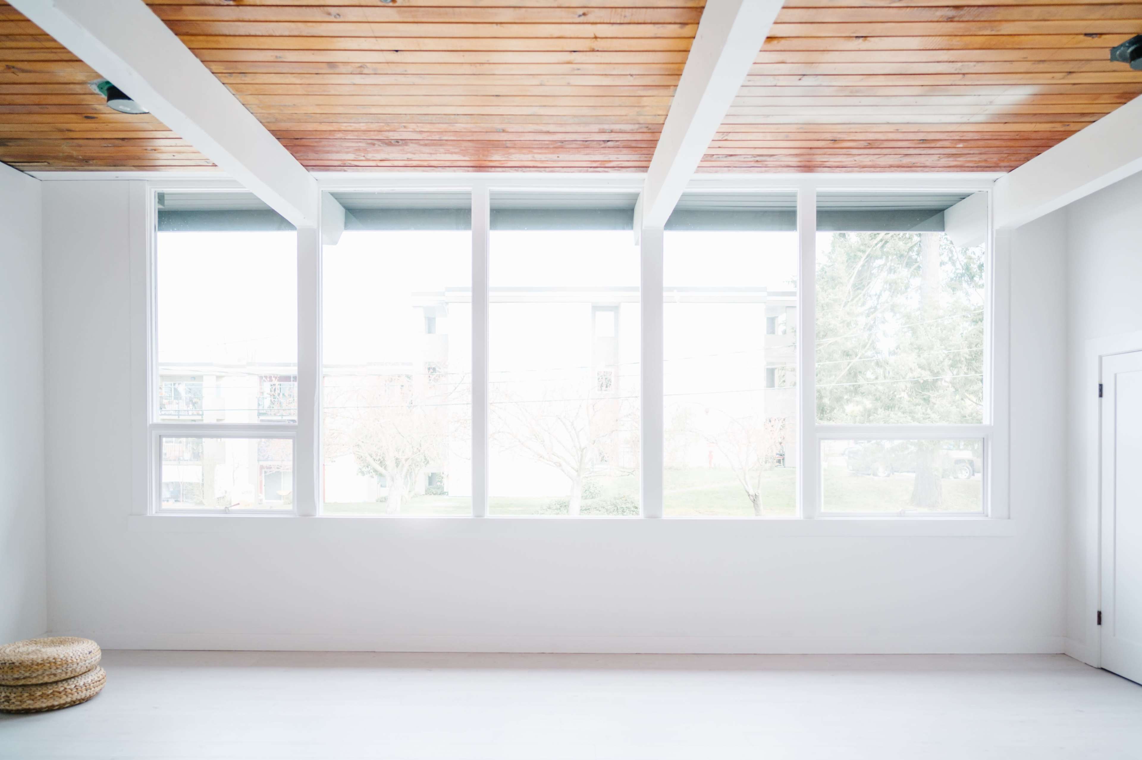 A minimalist interior room with large windows allowing natural light to fill the space, and a wooden ceiling above.