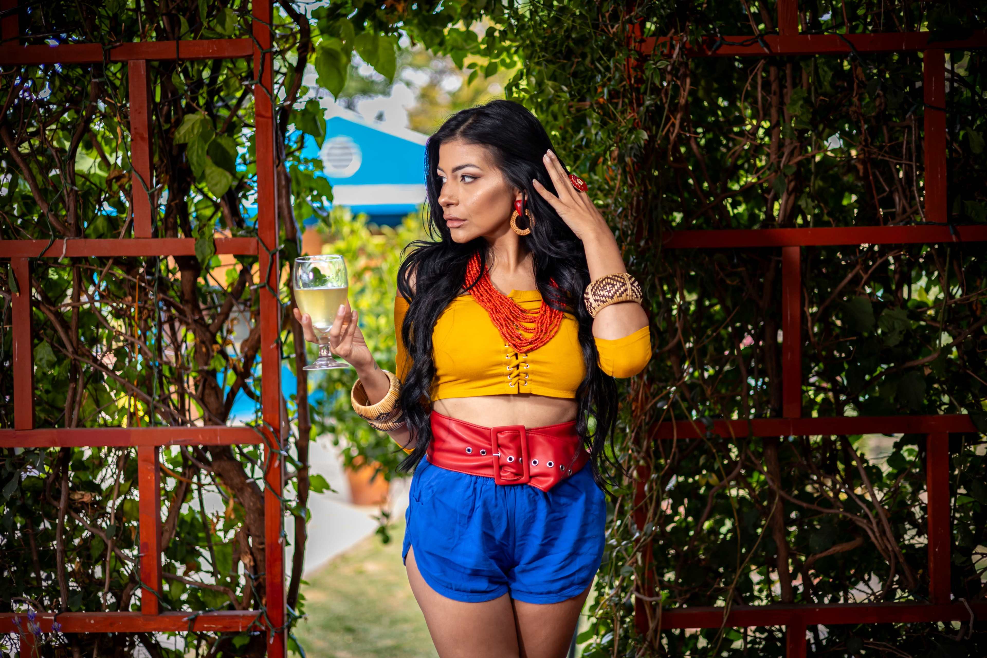 A woman wearing a colorful outfit stands in a garden, holding a drink and posing beside a trellis covered with greenery.