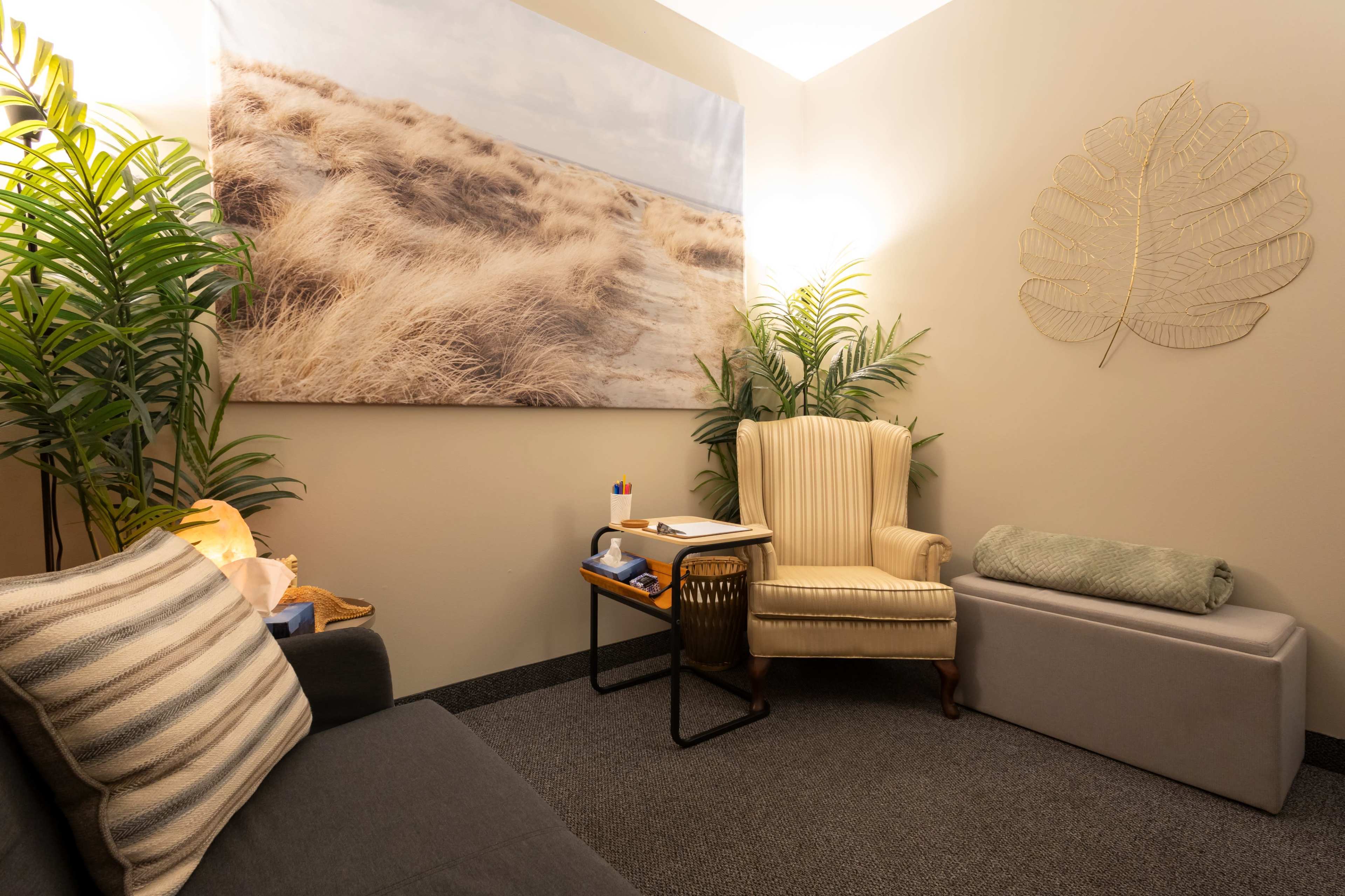 The image shows a cozy relaxation room with a beige armchair, a gray bench, a small table, and decorative plants, all oriented around a large beach-themed mural.