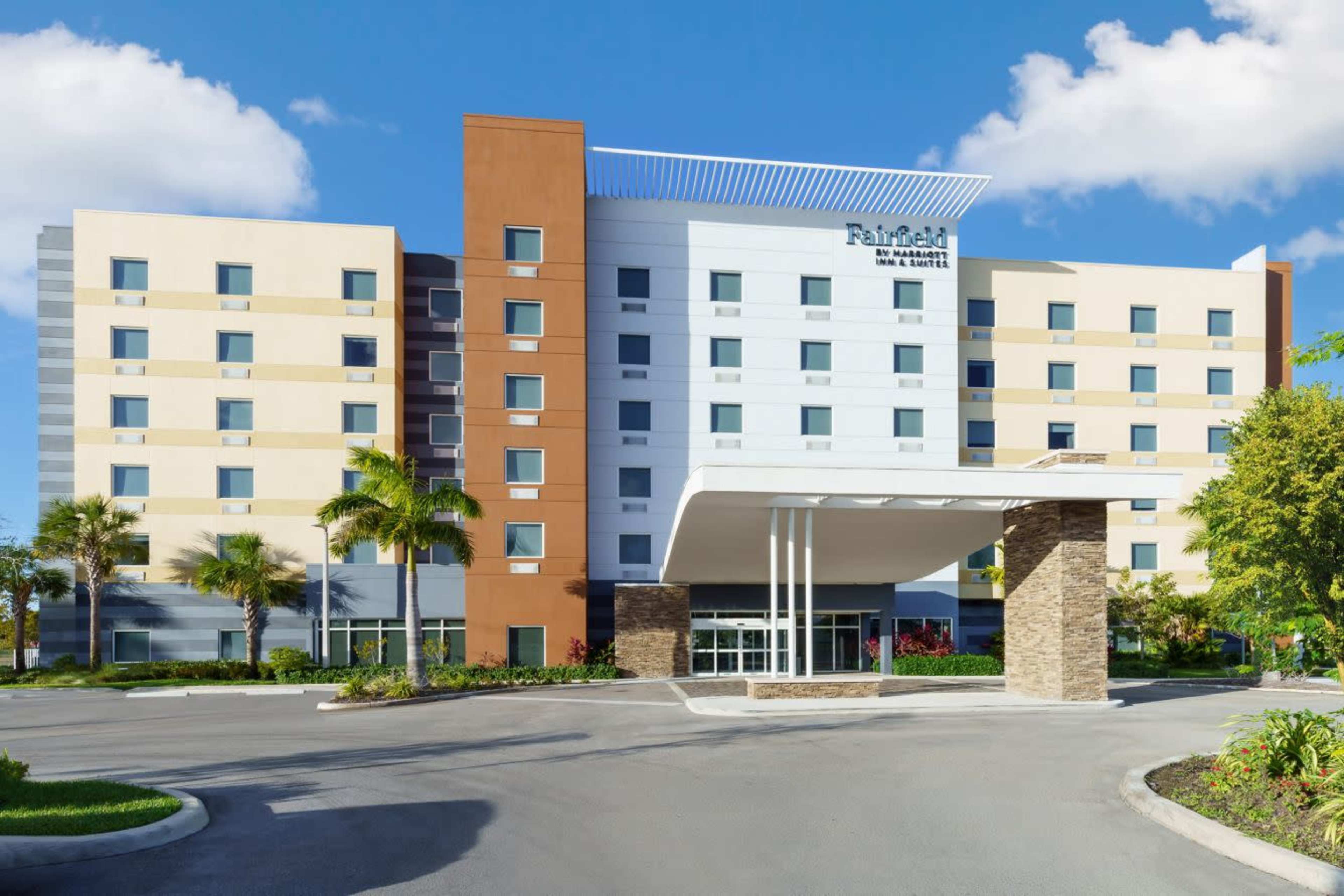 The image shows a modern hotel building with a multi-colored facade, featuring a covered entrance and landscaped surroundings.
