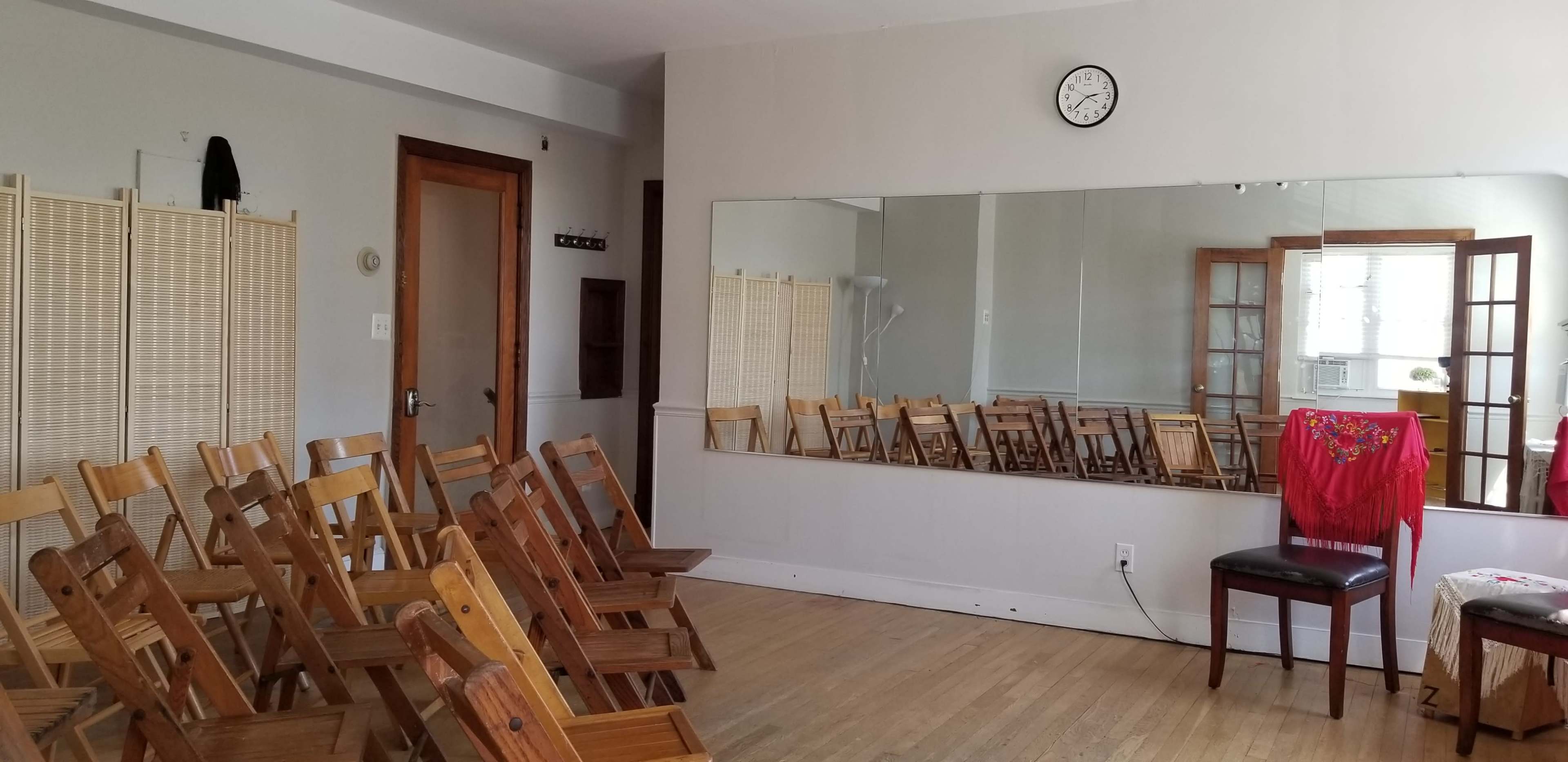 A spacious room with wooden folding chairs arranged in rows, a wall mirror reflecting the setup, and a clock on the wall.